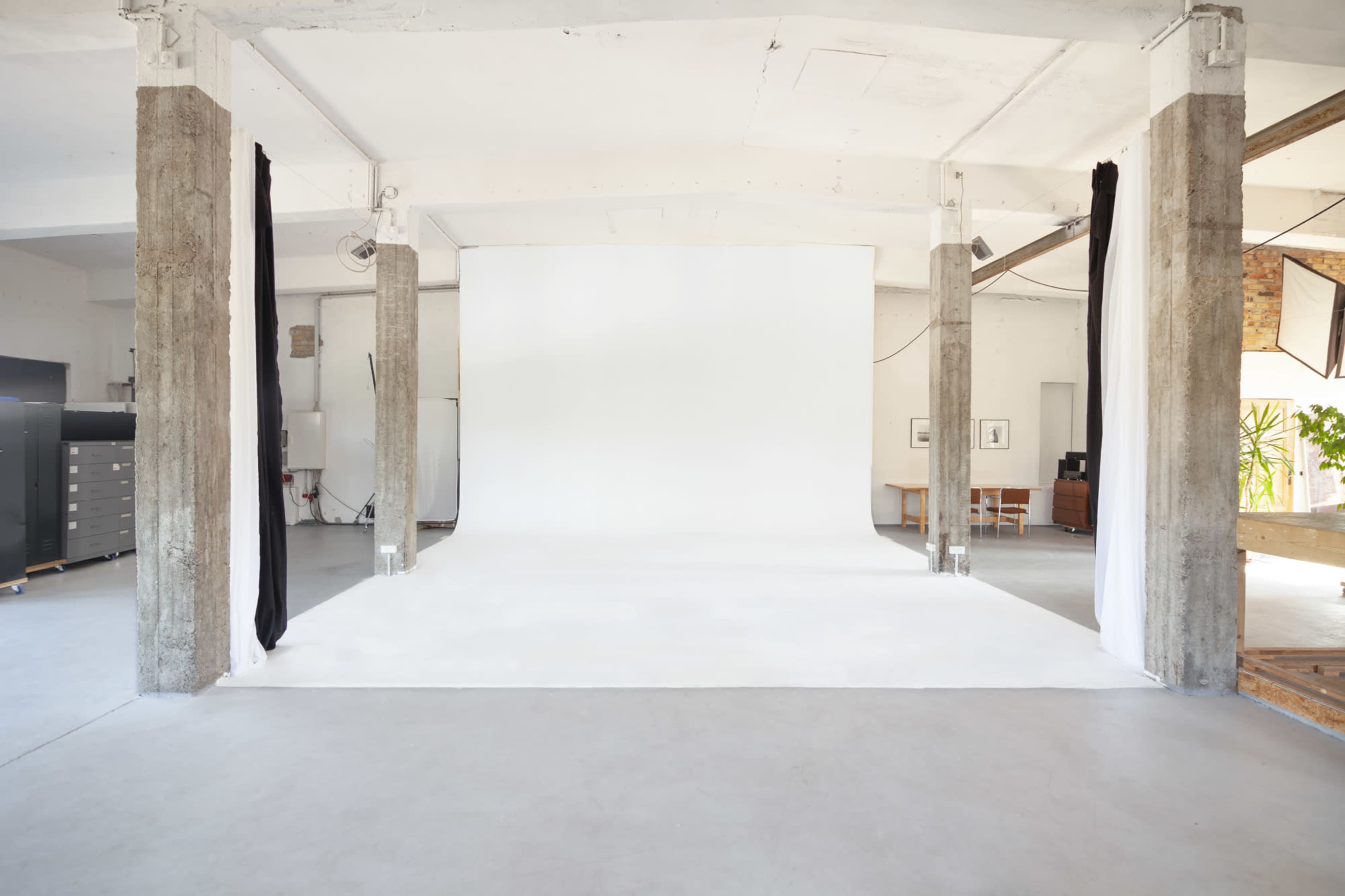 A spacious photography studio with a white backdrop and concrete pillars, featuring a minimalist design and neutral colors.