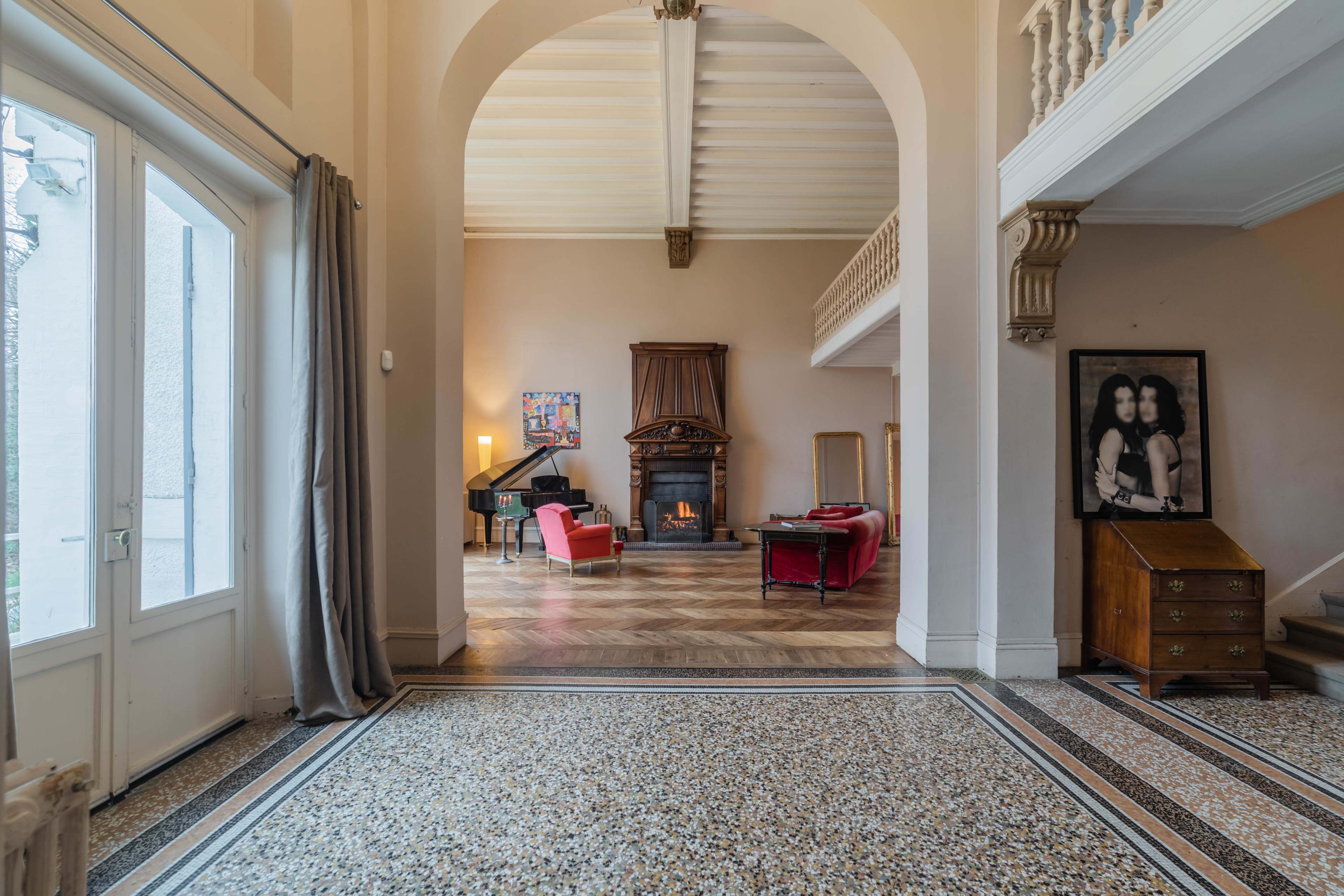 A spacious interior with a fireplace, a grand piano, and furniture arranged in a well-lit living area featuring intricate flooring and large windows.
