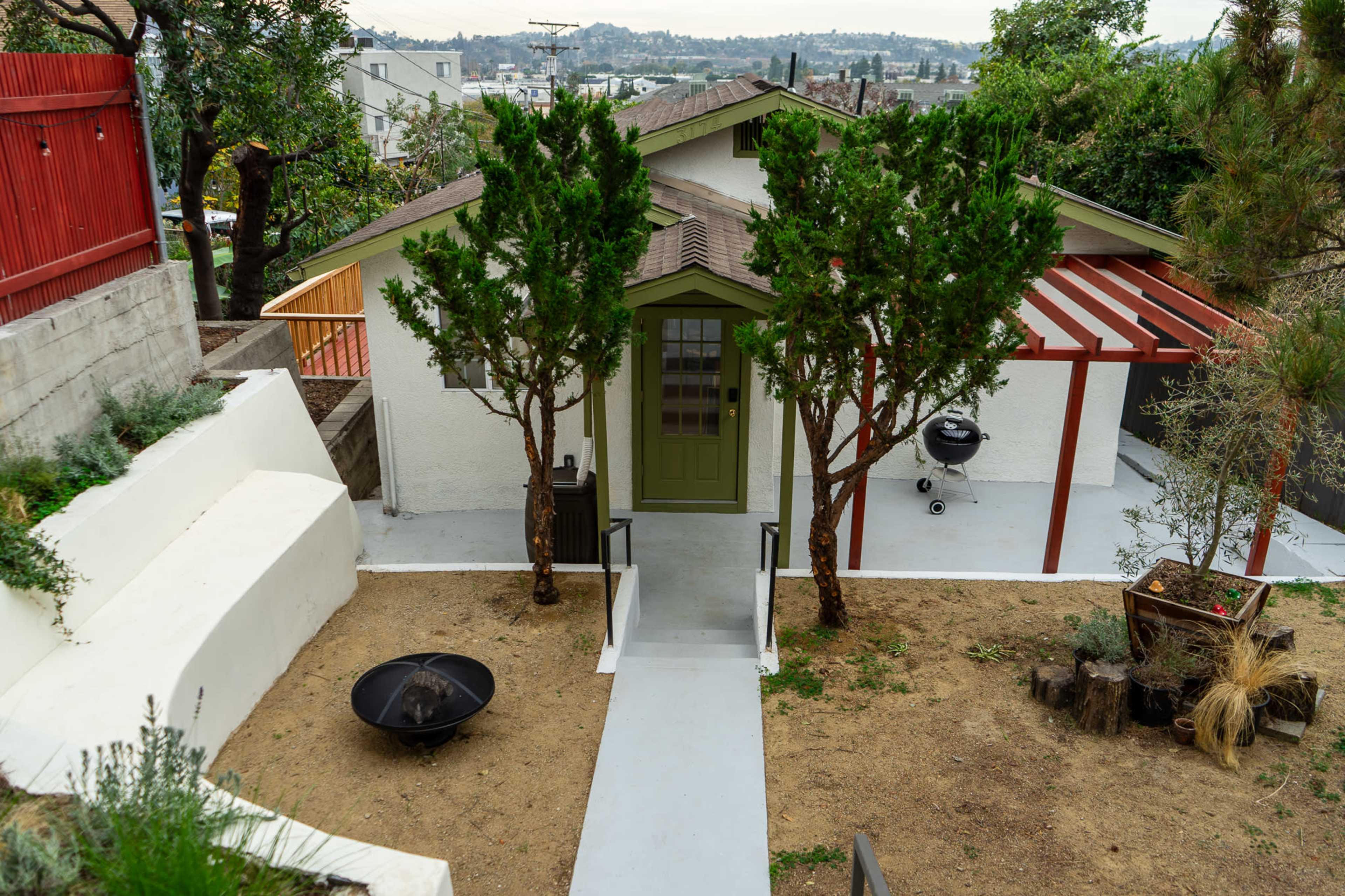 The image shows a house with a green door surrounded by two trees, a gravel area, and a small grilling station under a red pergola.