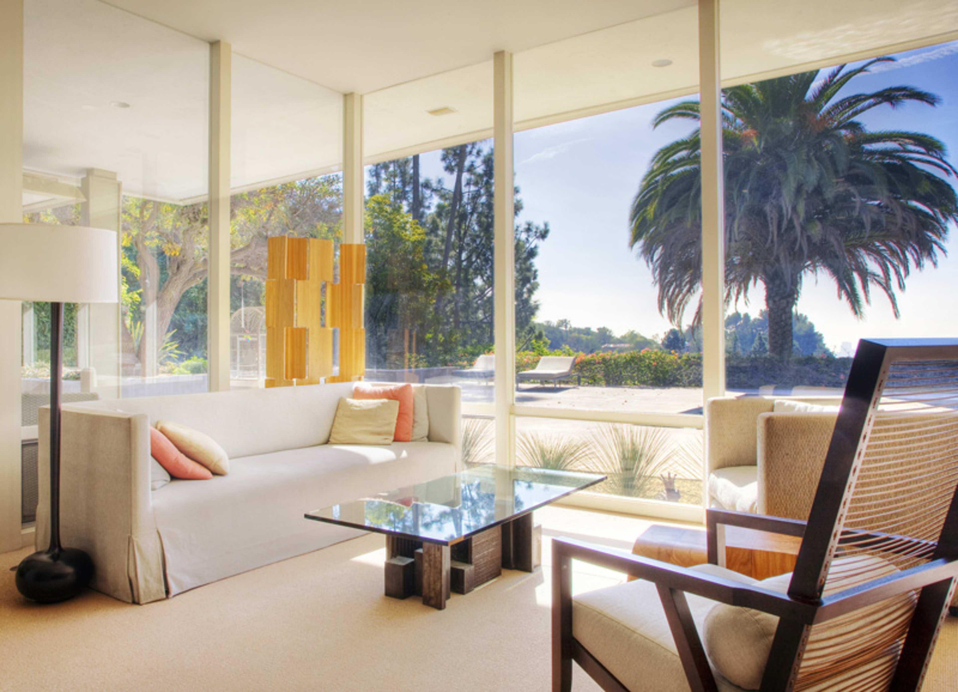 A modern living room features a light-colored sofa, a glass coffee table, and large windows that overlook a garden with palm trees.