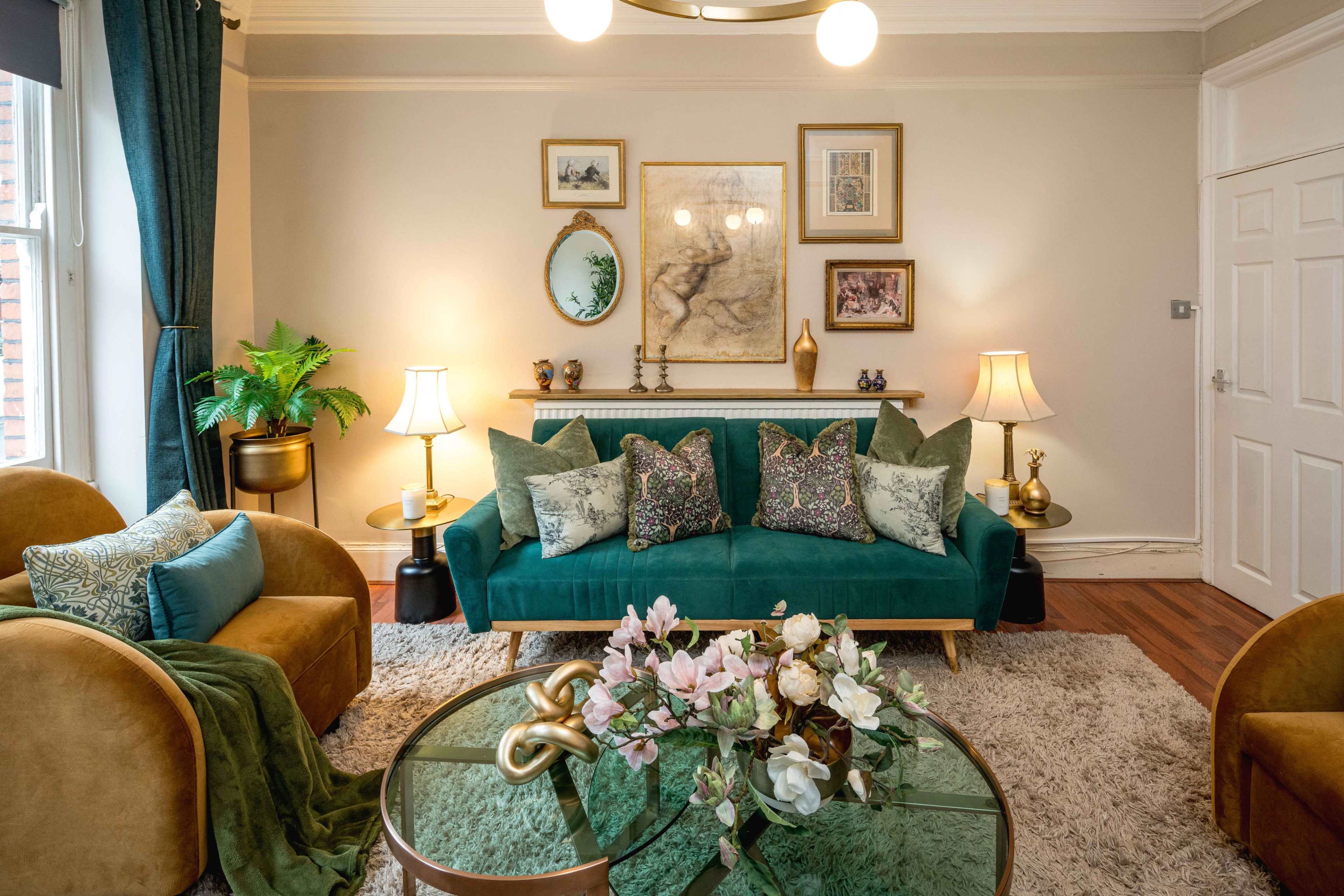 The image showcases a stylish living room featuring a teal sofa with decorative cushions, two golden accent chairs, a round glass coffee table adorned with flowers, and various framed artworks on the walls.