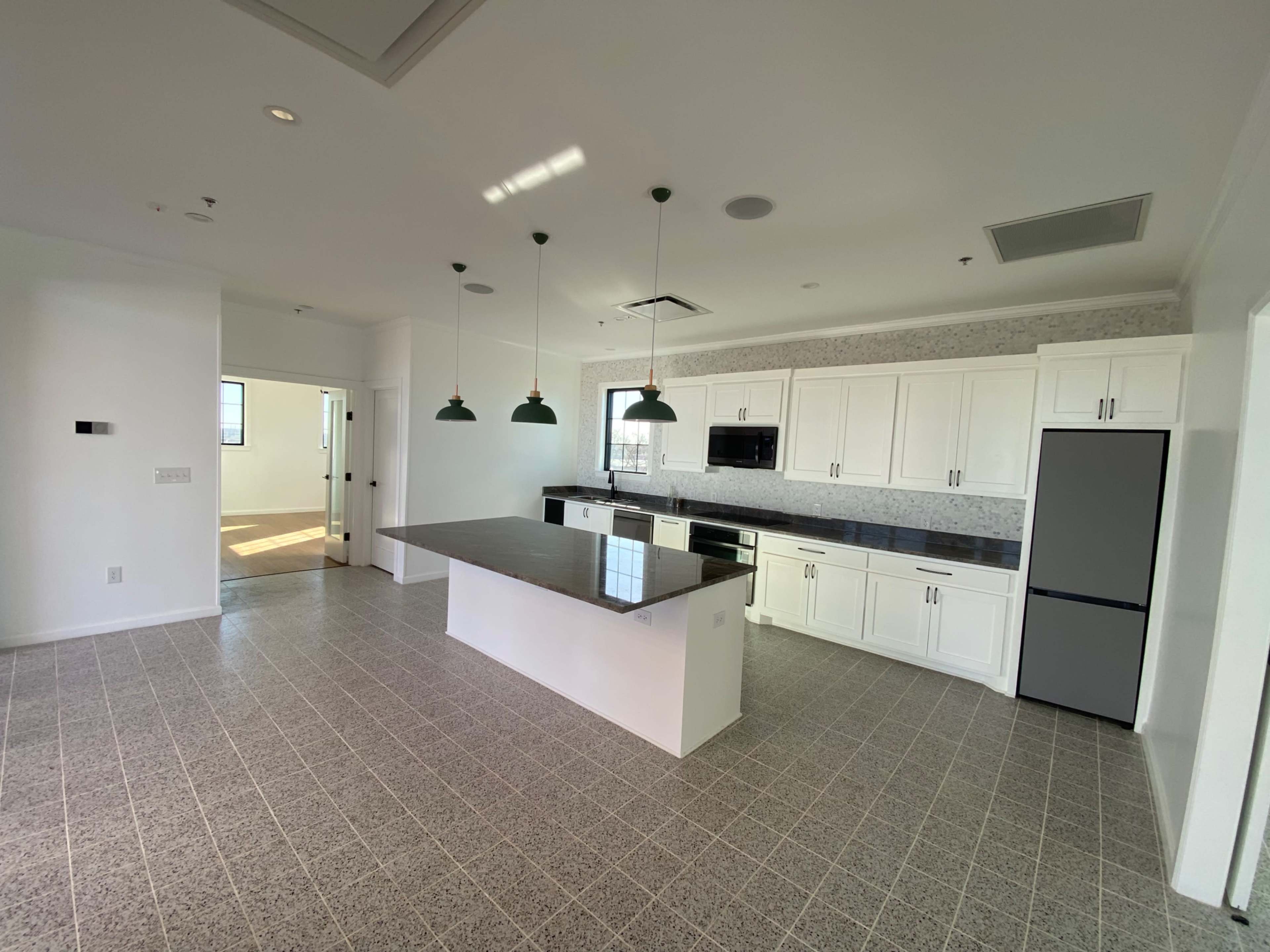 A modern kitchen with white cabinets, a dark countertop, and pendant lights, adjacent to a connected open space.