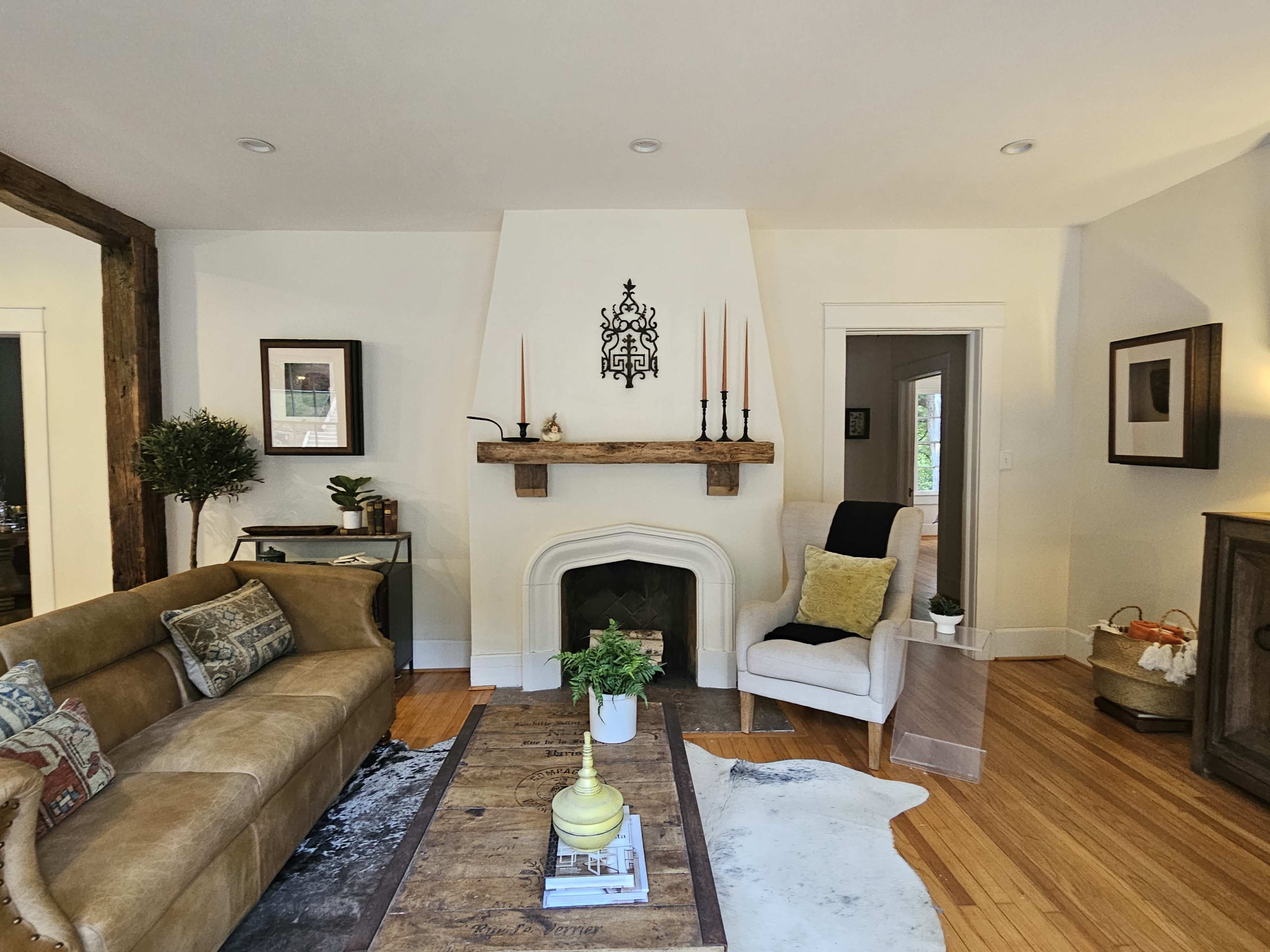 A cozy living room features a brown sofa, a wooden coffee table, and a white fireplace flanked by candles and plants.