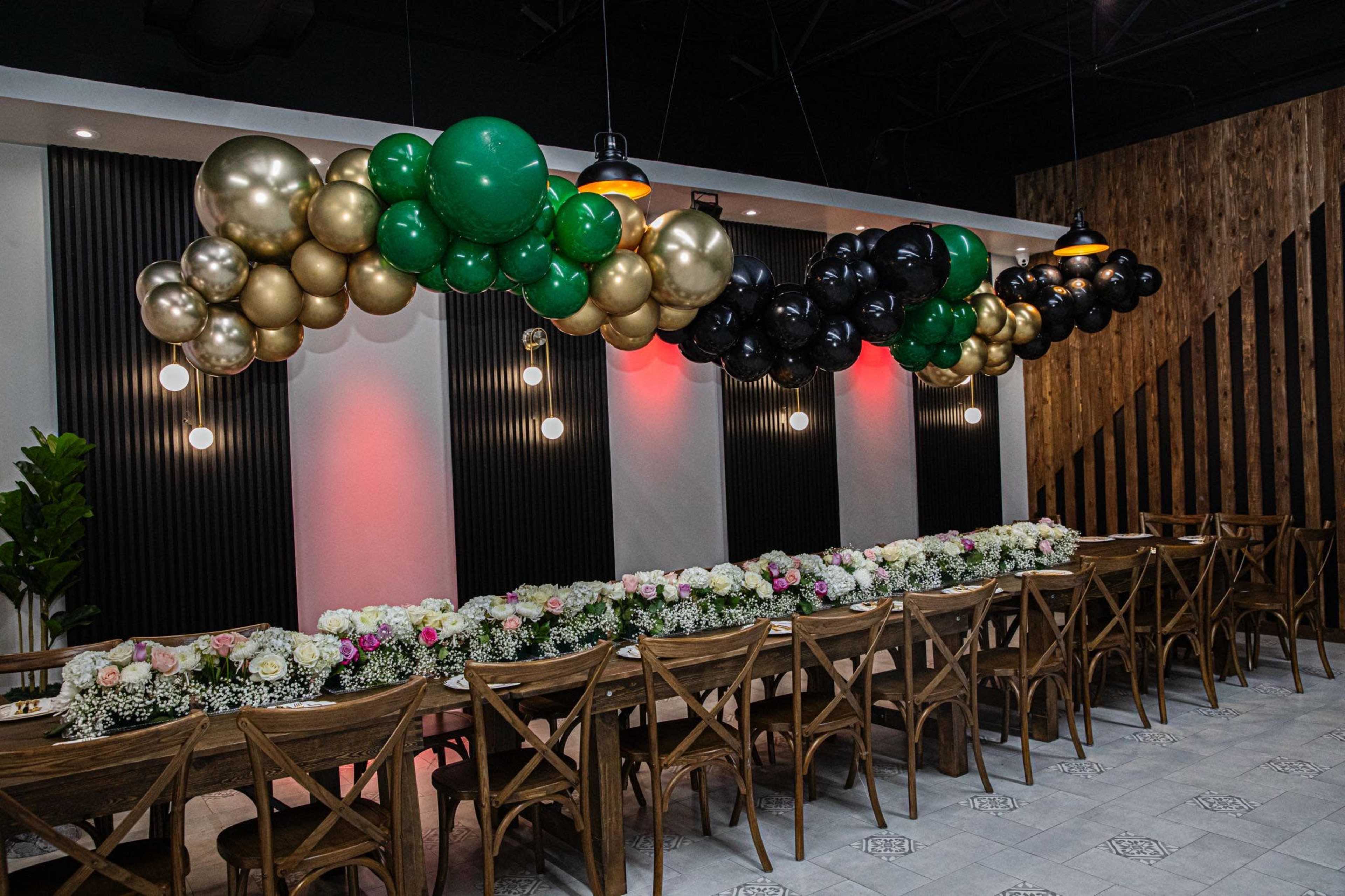 The image shows a dining area decorated with a balloon arch in black, gold, and green above a long table adorned with floral arrangements and wooden chairs.
