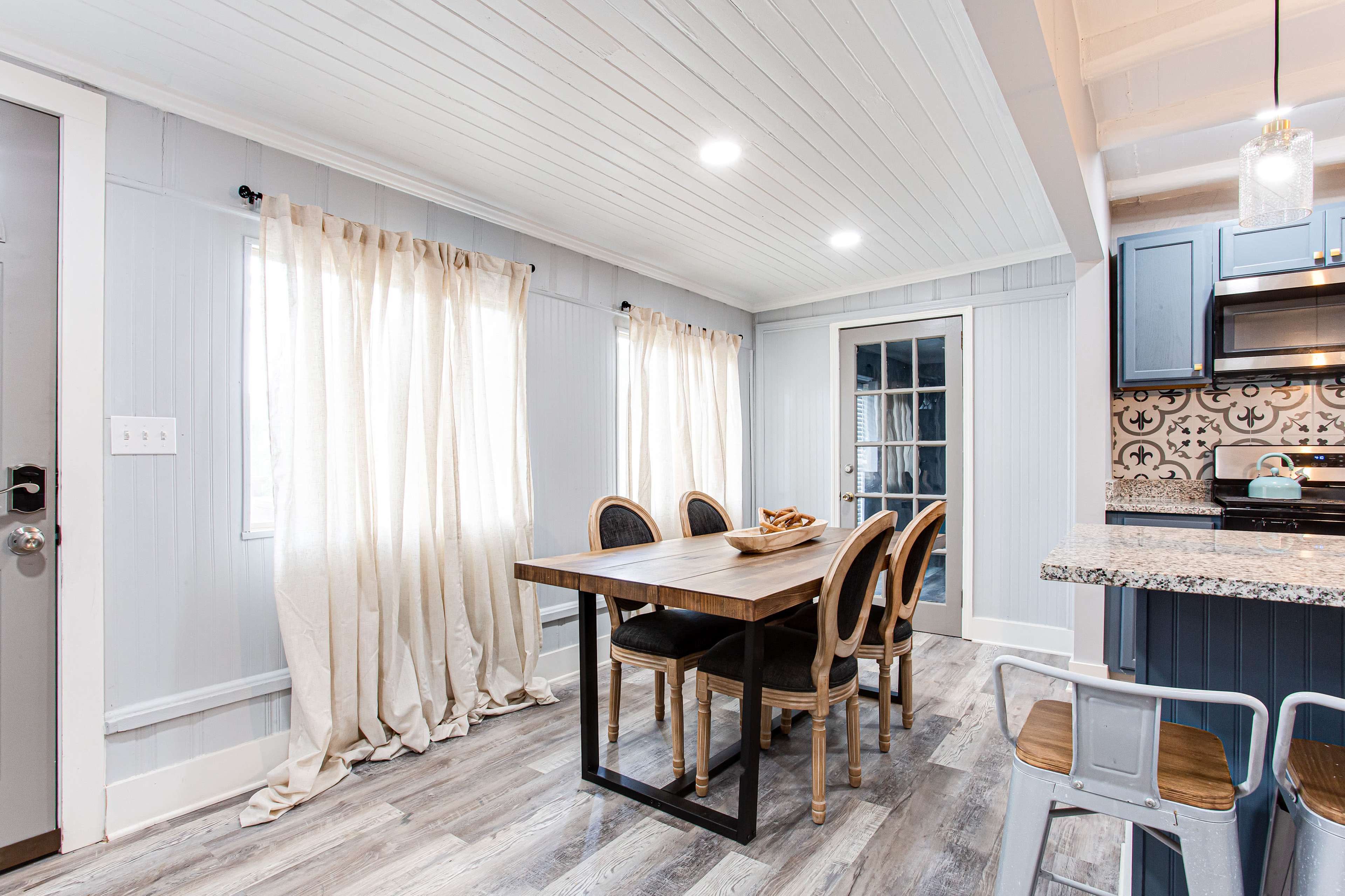 A dining area features a wooden table surrounded by chairs, with large windows dressed in light curtains and a door leading to a patio.