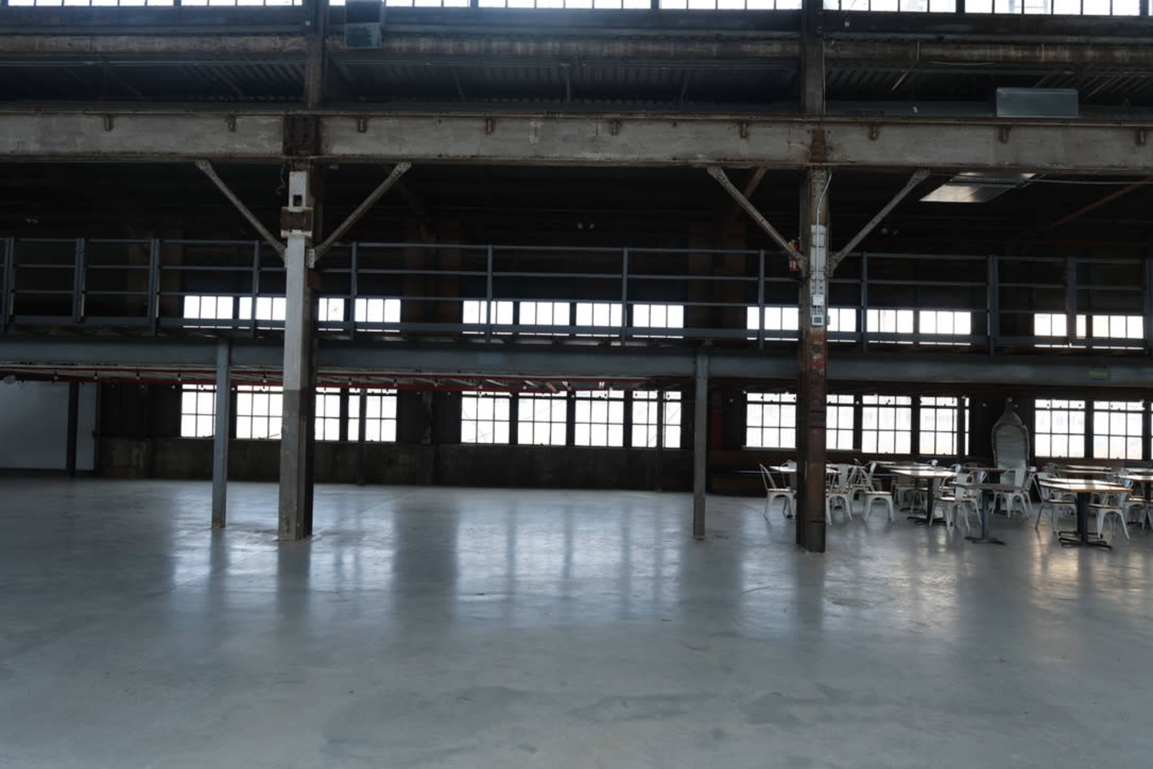 The image shows an empty industrial-style interior with large windows and metal columns, featuring a sparse arrangement of tables and chairs.