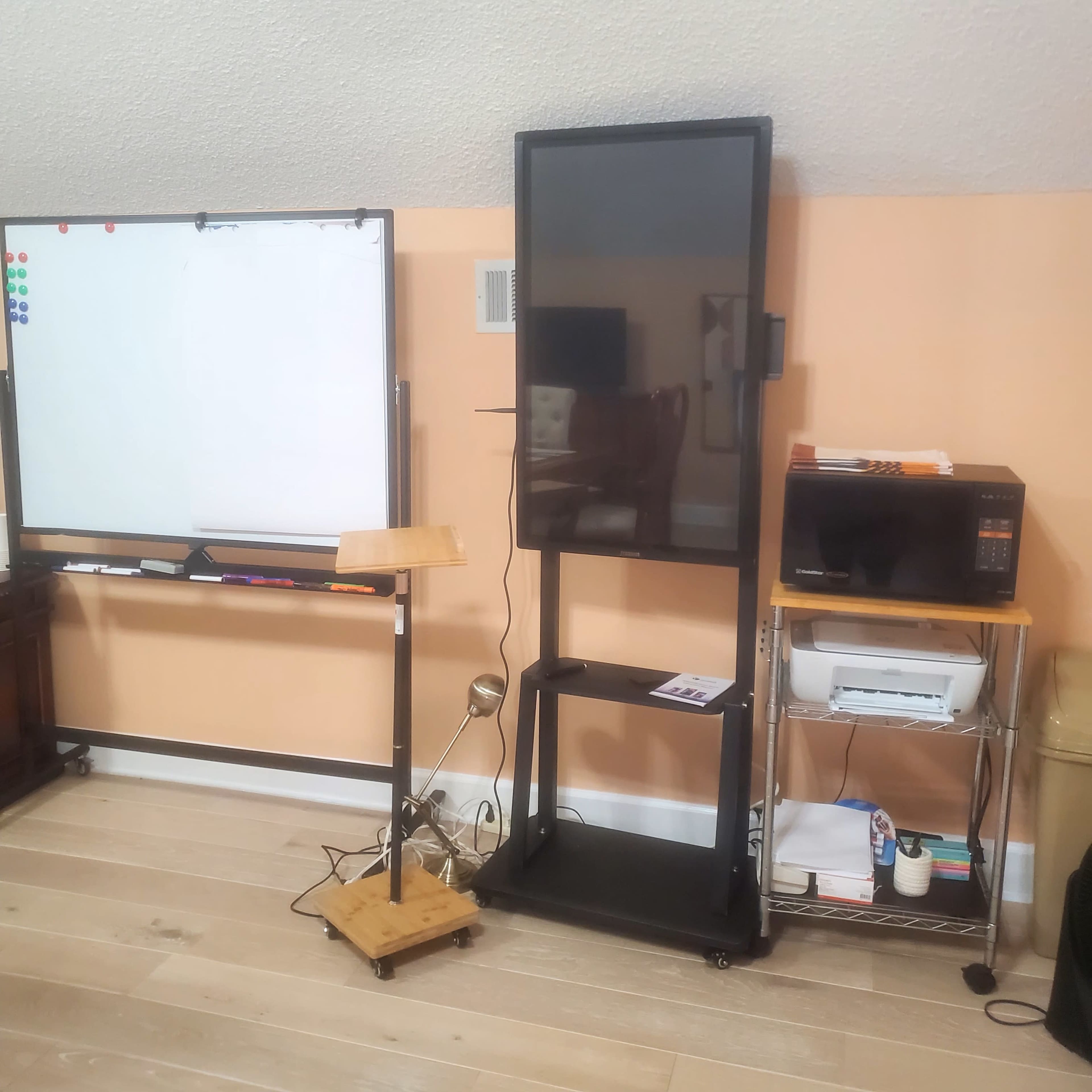 A room with a mobile TV stand holding a television, a whiteboard, a microwave, and a small wooden table next to them.