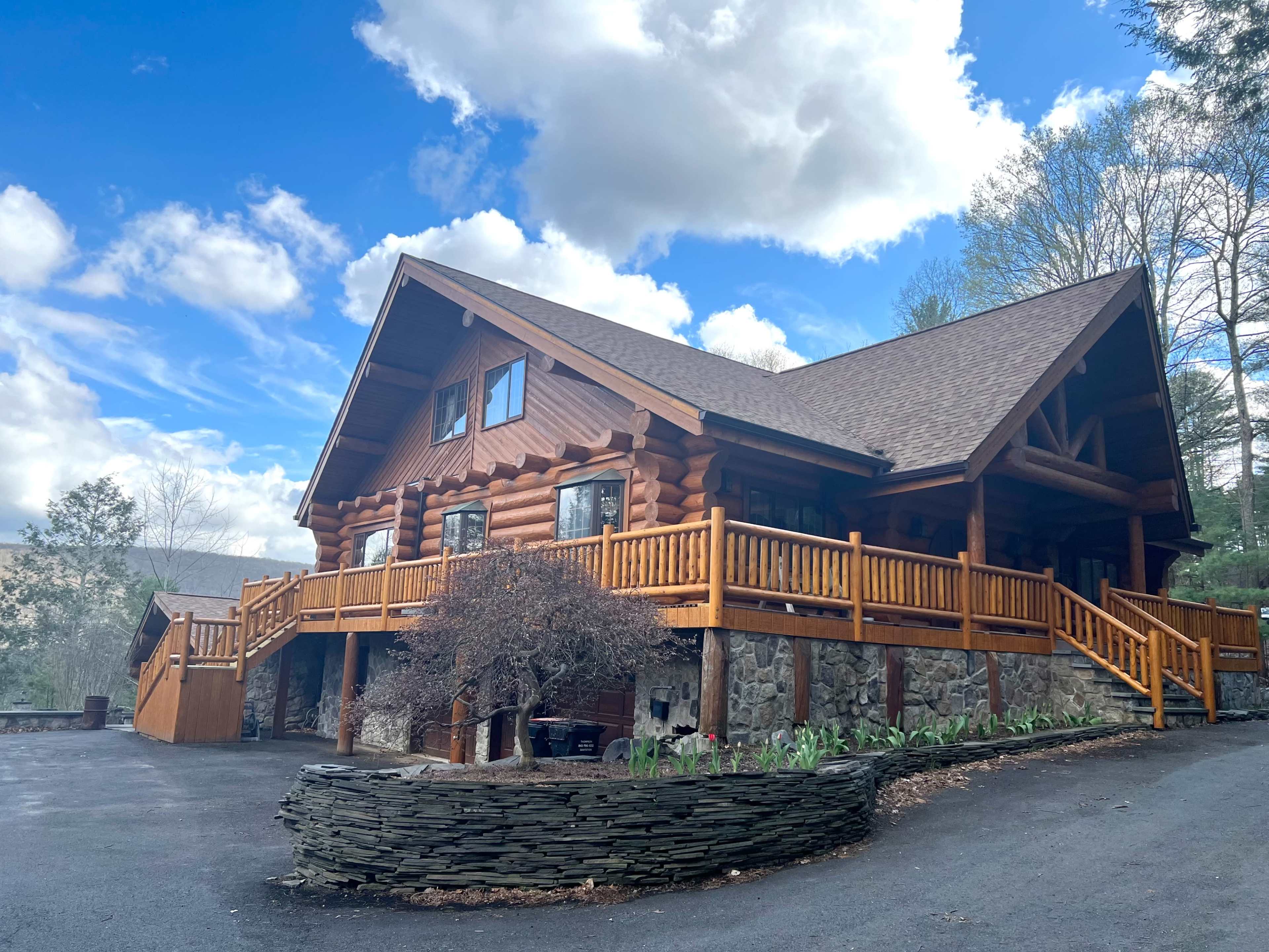 A large log cabin with a wraparound porch is set against a backdrop of trees and a blue sky with clouds.