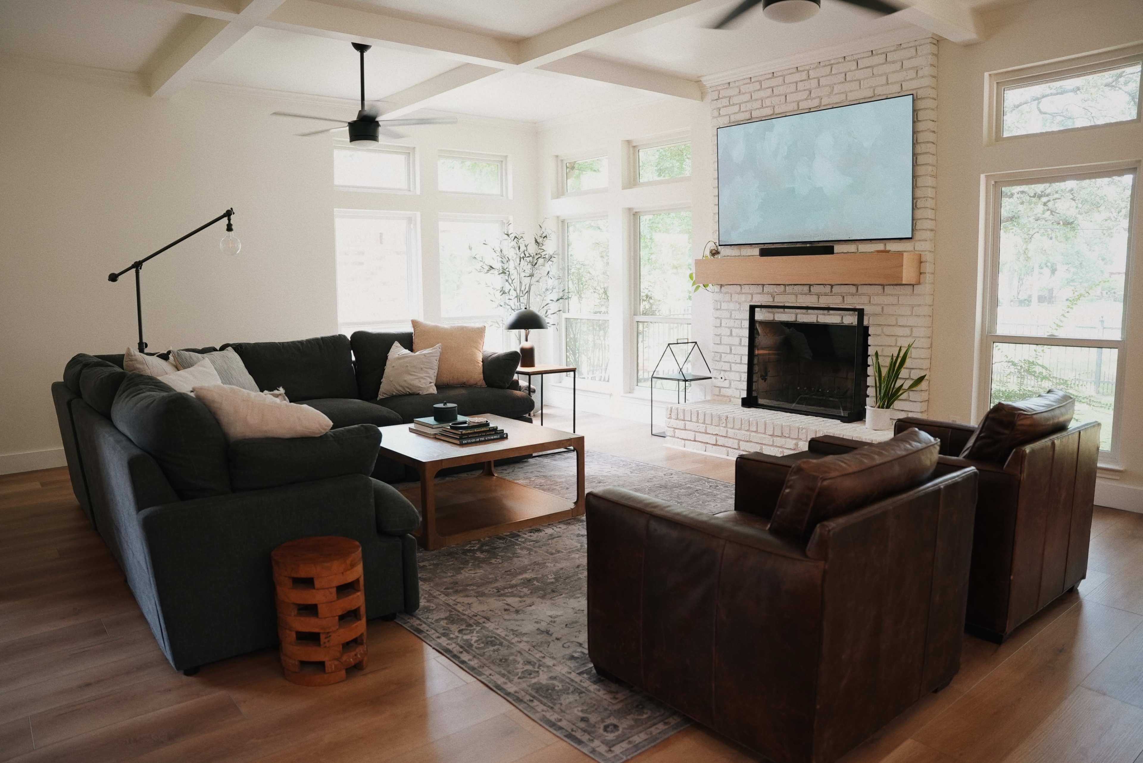 The image shows a modern living room with a large sectional sofa, two leather armchairs, a wooden coffee table, a brick fireplace, and a flat-screen TV mounted on the wall.