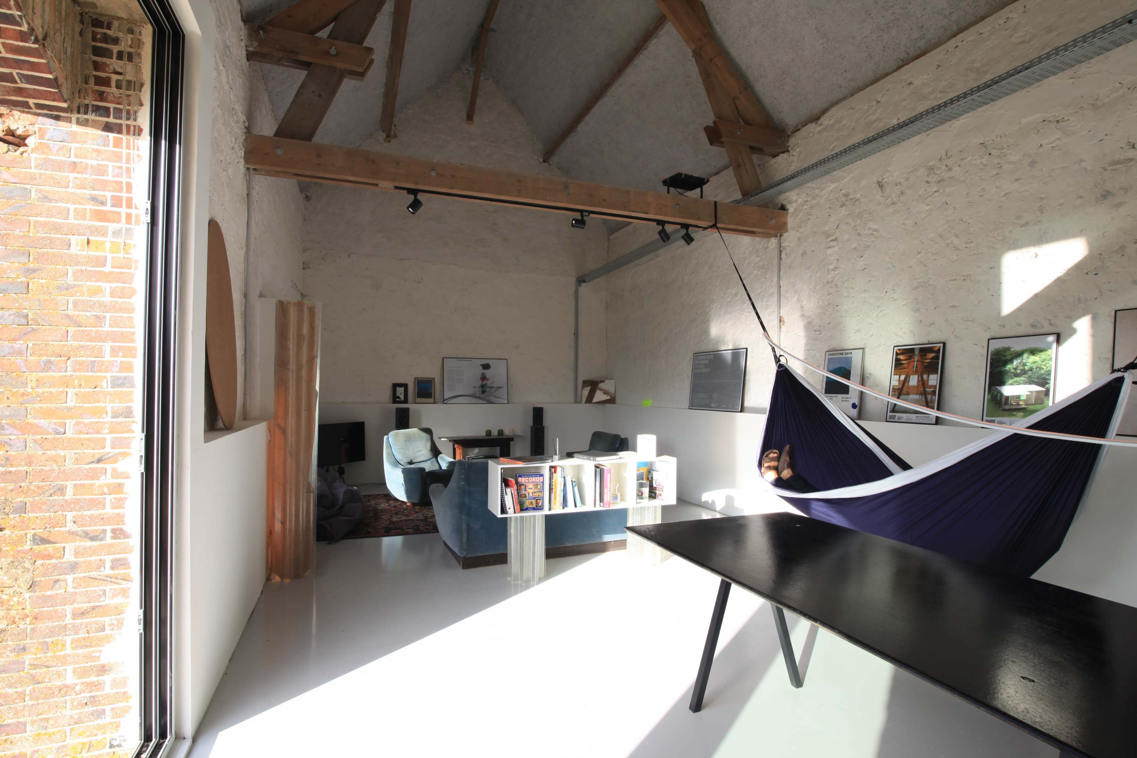The image shows a spacious room with high ceilings, featuring a hammock, a dark table, blue chairs, and various decorations on the walls.