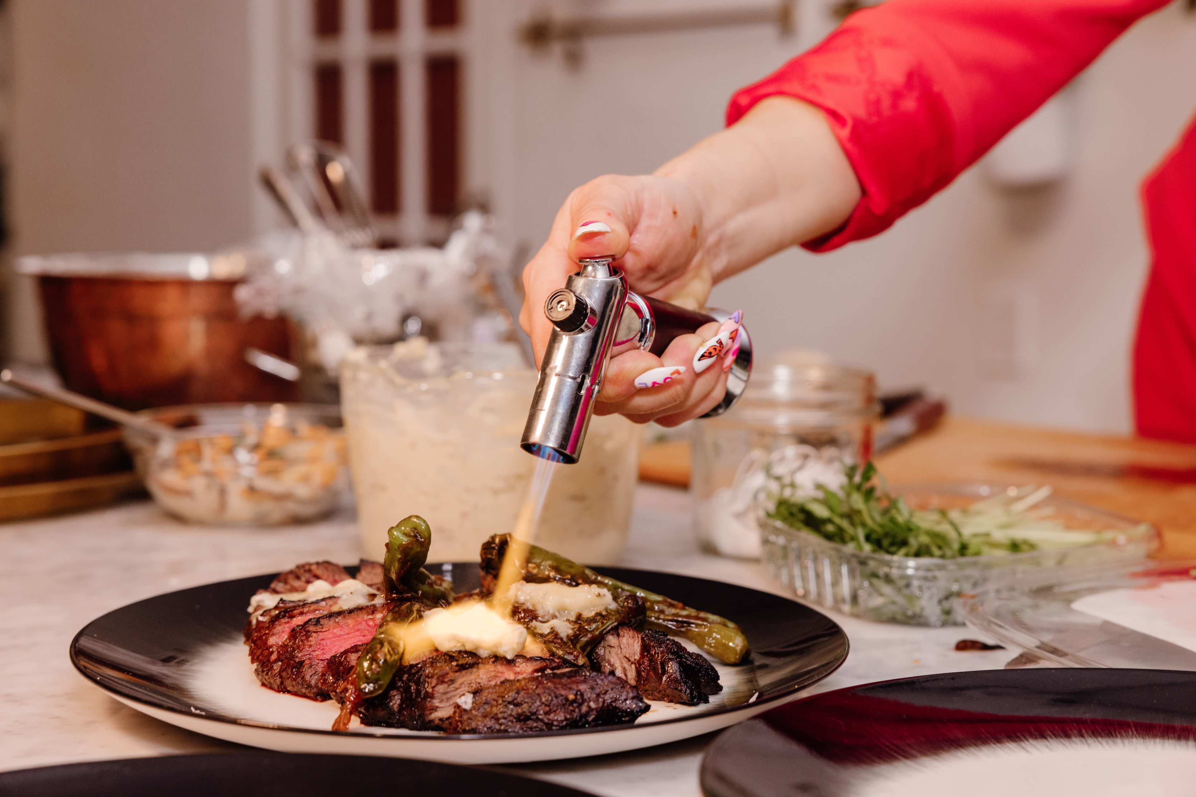 A person is using a culinary torch to caramelize the top of a plated dish featuring steak and asparagus in a kitchen setting.