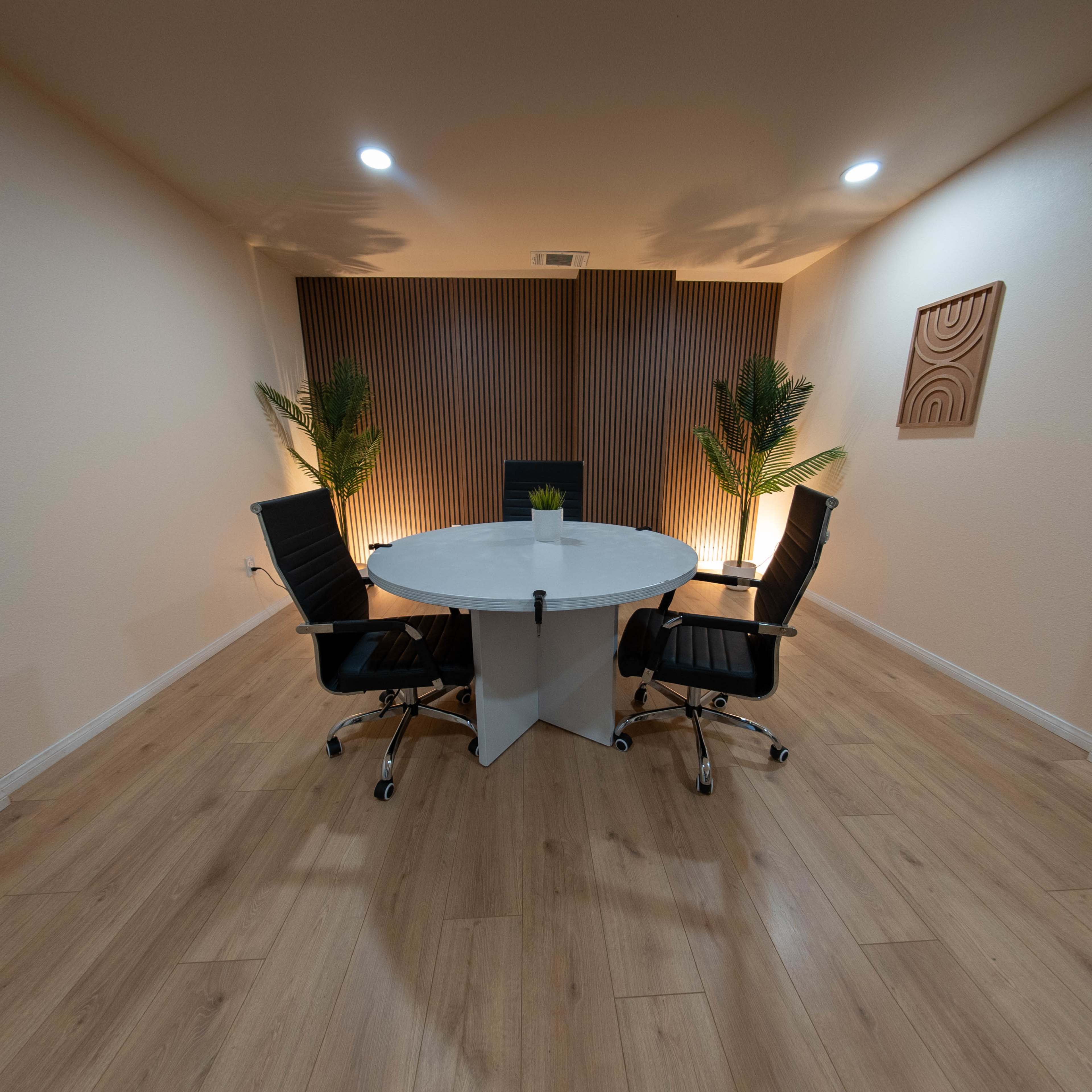 A modern conference room features a circular white table surrounded by four black chairs, with wooden paneling and potted plants in the background.