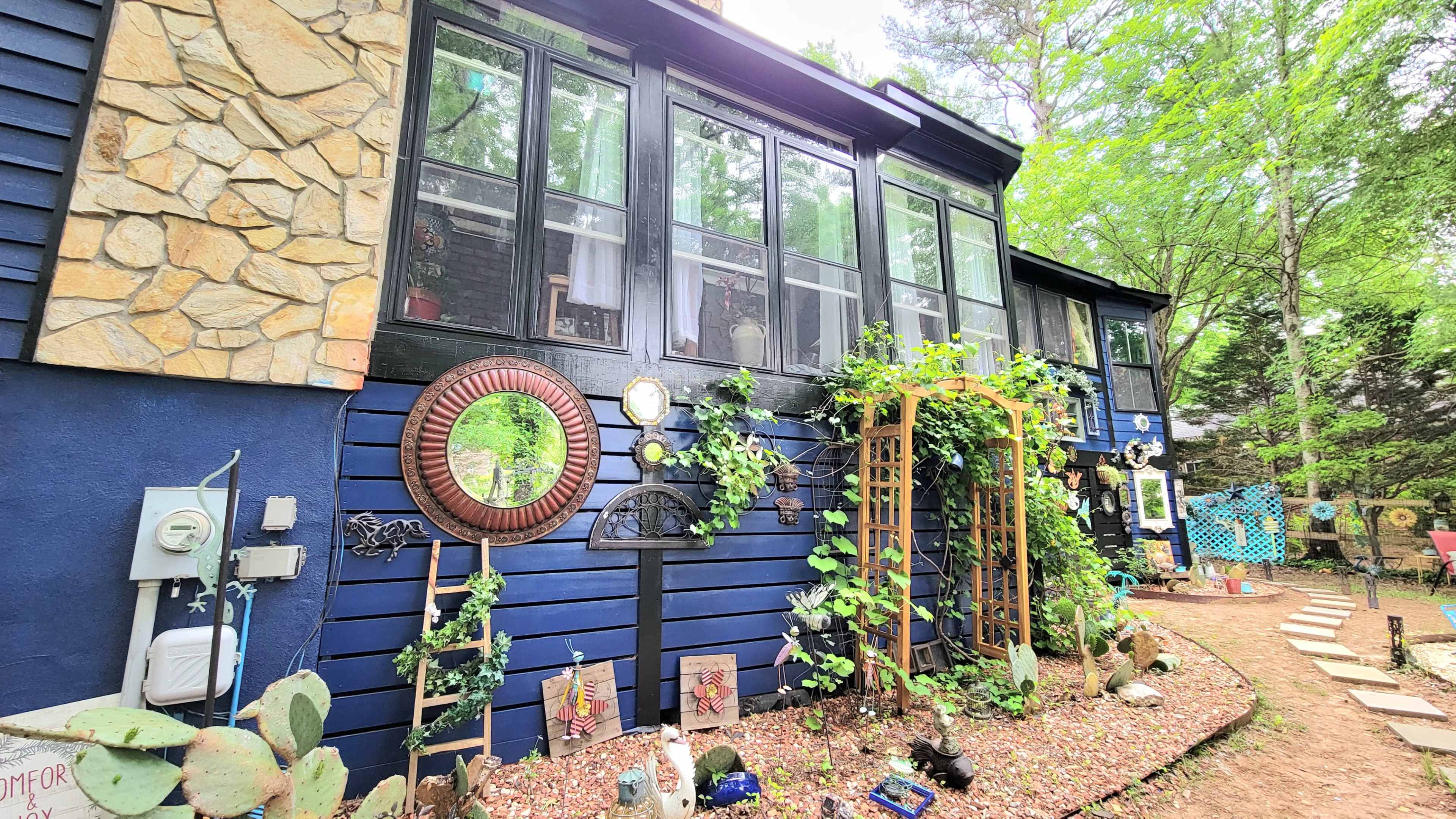 A blue house with large windows, surrounded by greenery and a garden featuring decorative elements like a mirror and trellis.