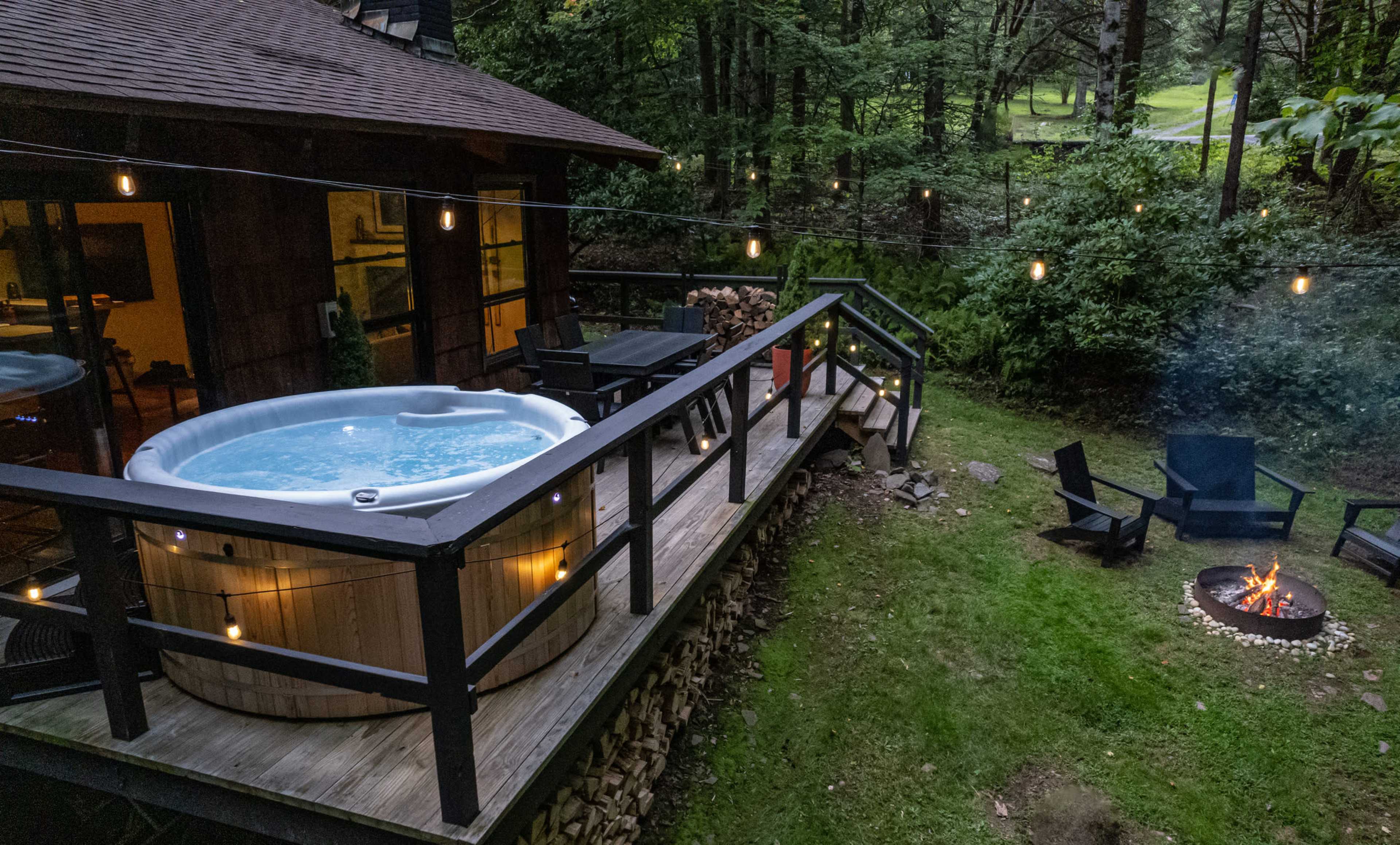 A hot tub is located on a wooden deck next to a fire pit surrounded by chairs, set in a wooded area.