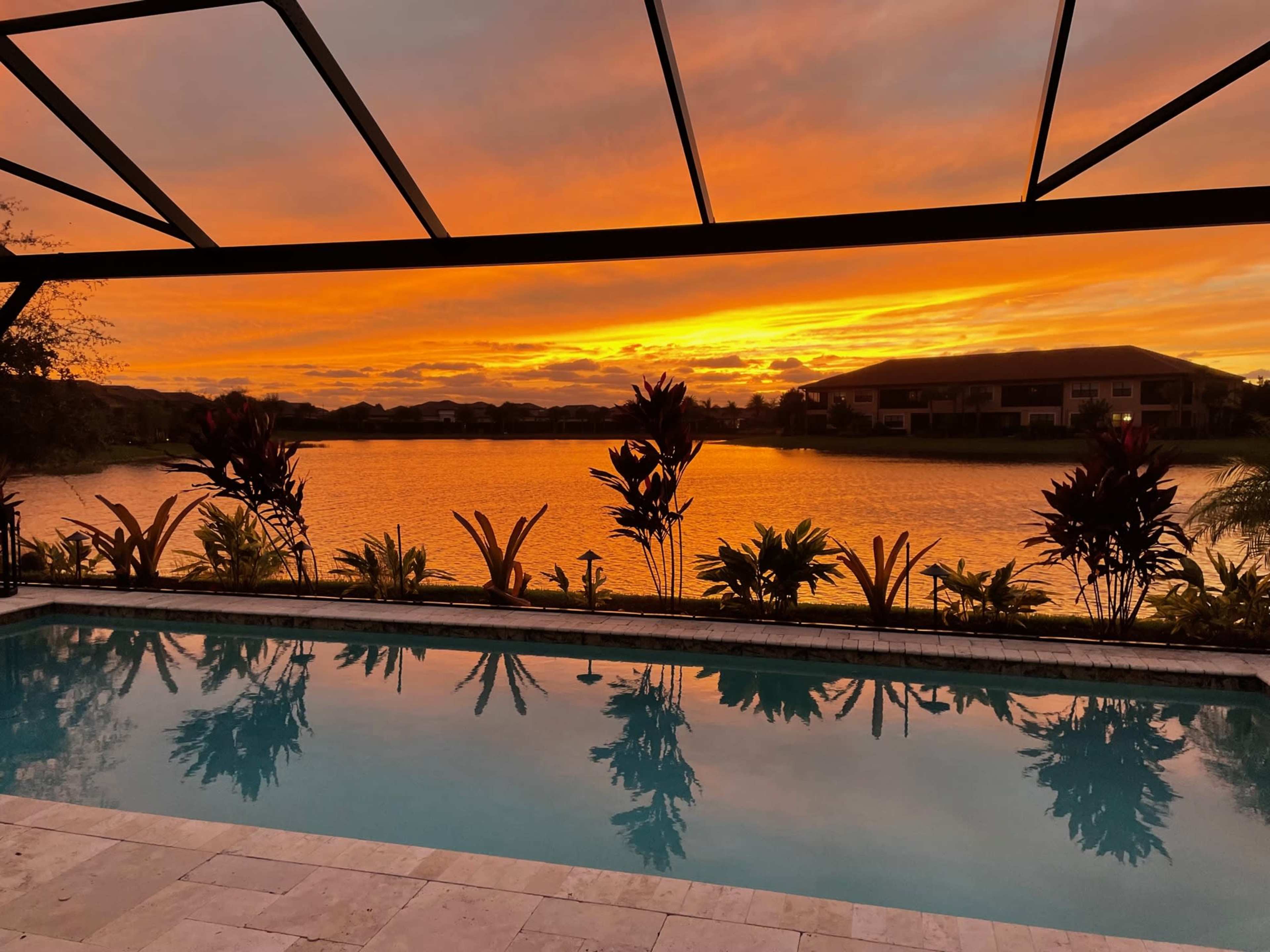 A pool surrounded by plants, with a vibrant sunset reflected in the water and a lake beyond.