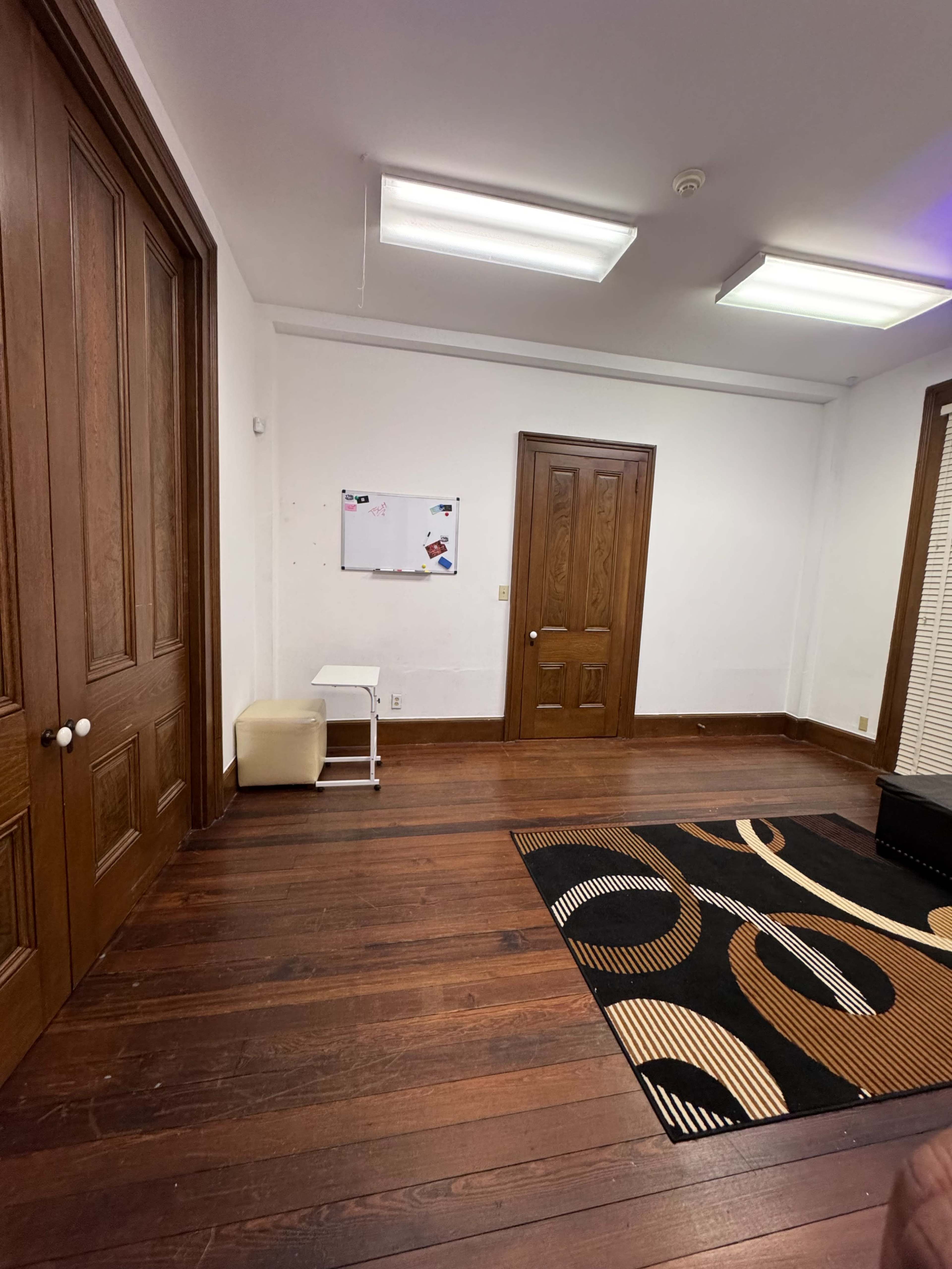 The image shows a sparse room with wooden floors, a whiteboard on the wall, a closed door, and a black rug featuring a circular design.