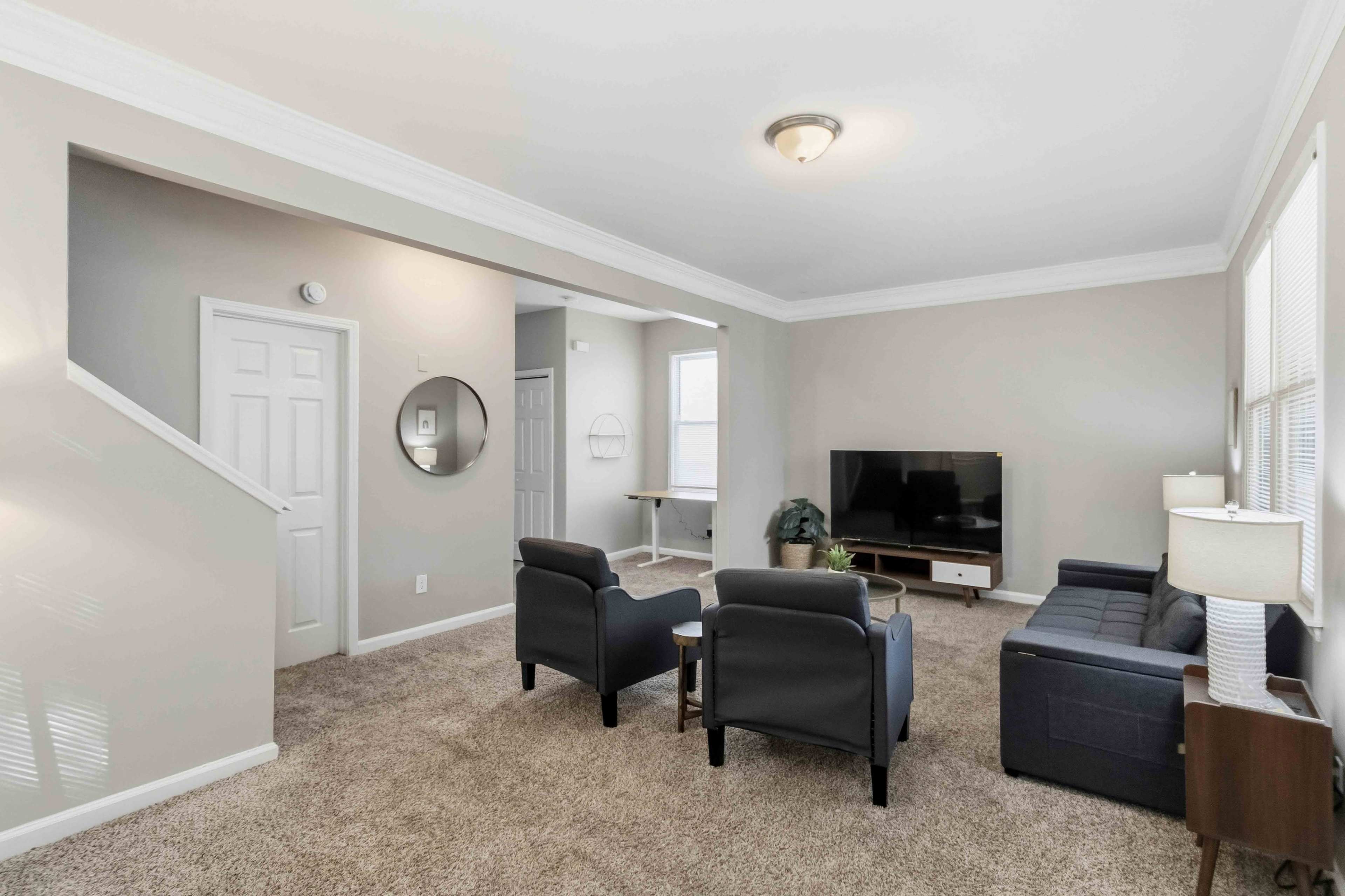 The living room features a comfortable seating arrangement with two armchairs and a sofa, a television mounted on a stand, and a staircase in the corner.