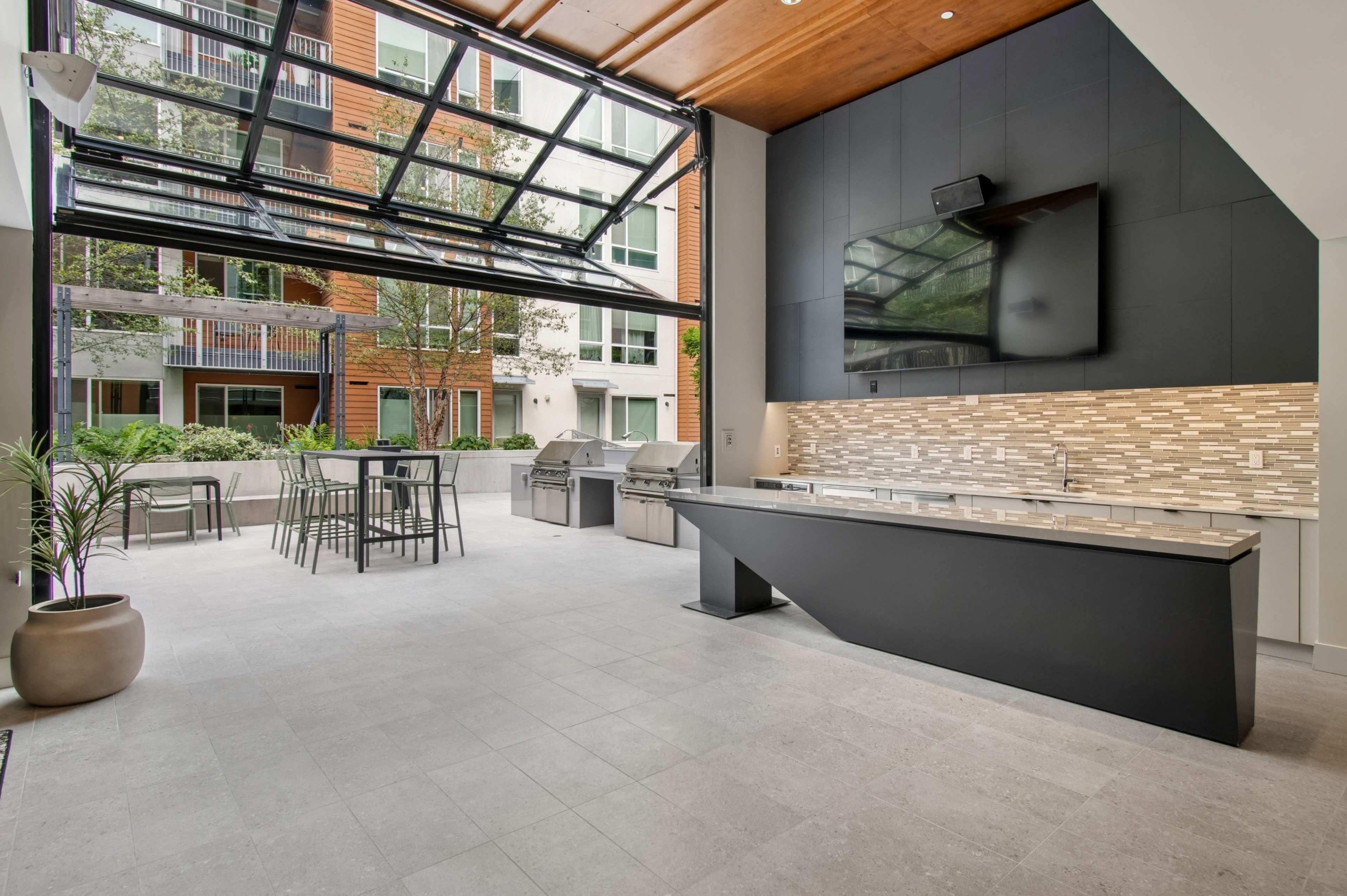 The image depicts a modern outdoor kitchen area featuring a spacious layout with a large island, grill, seating for several people, and large glass doors that open to a green outdoor space.