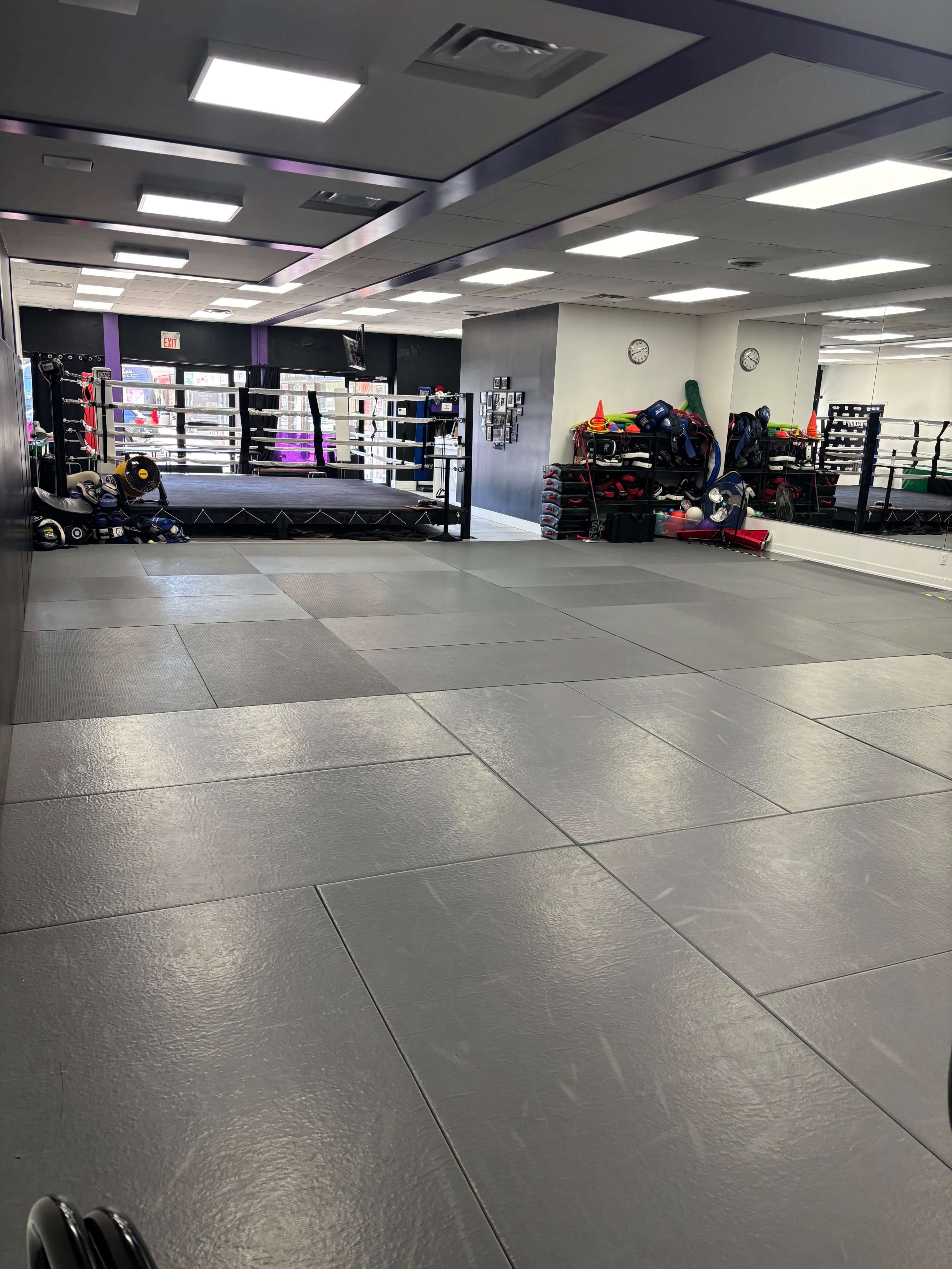 The image shows an empty training facility with gray flooring, training equipment stacked against the walls, and a boxing ring in the background.