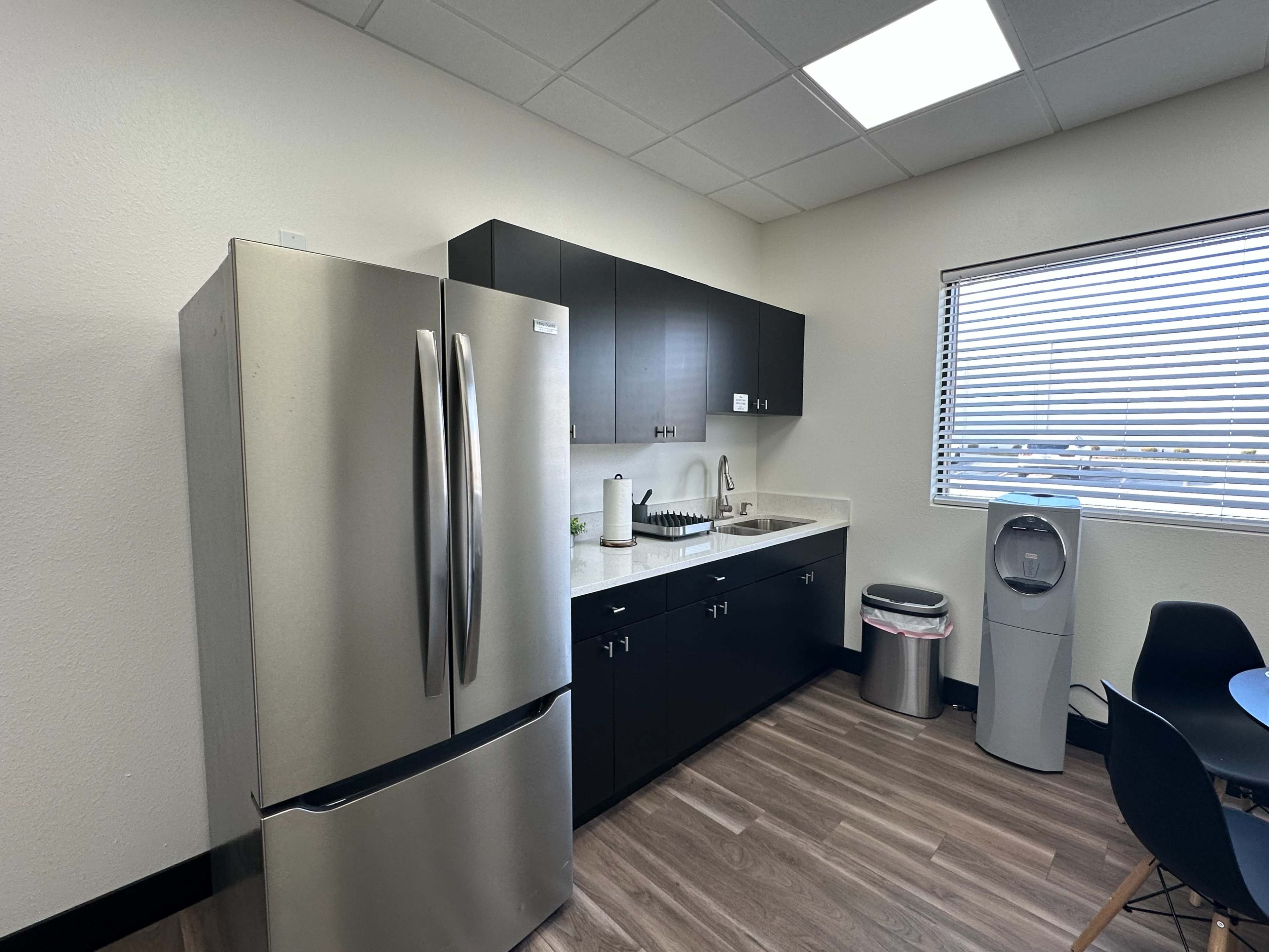 The image shows a modern kitchen area with stainless steel appliances, dark cabinets, and a countertop, featuring a sink and a water dispenser.