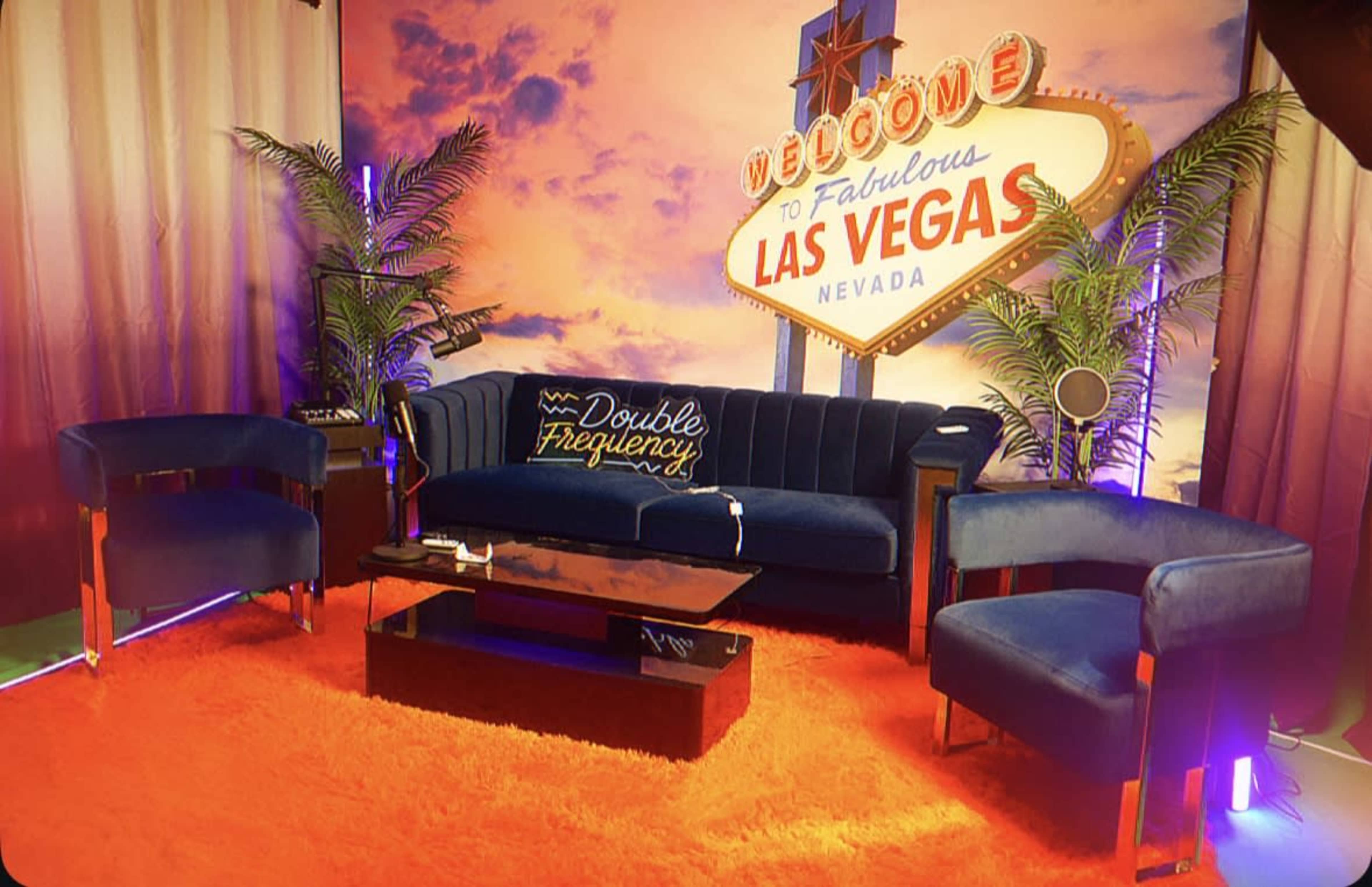 The scene features a blue sofa and matching chairs arranged around a black coffee table on an orange rug, set against a backdrop with a neon-lit "Welcome to Fabulous Las Vegas" sign.