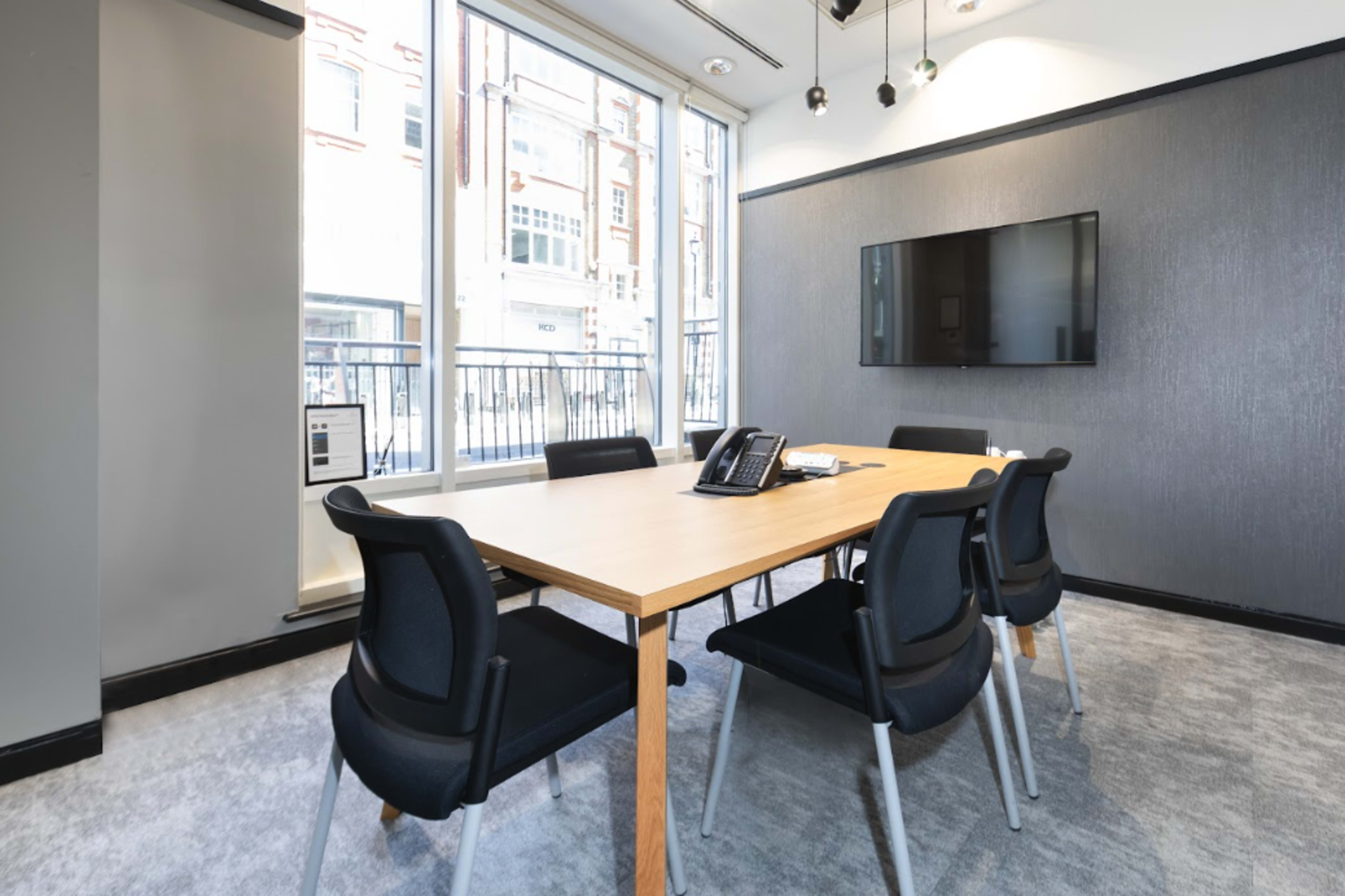 The image shows a modern conference room with a wooden table surrounded by black chairs, a wall-mounted TV, and large windows letting in natural light.