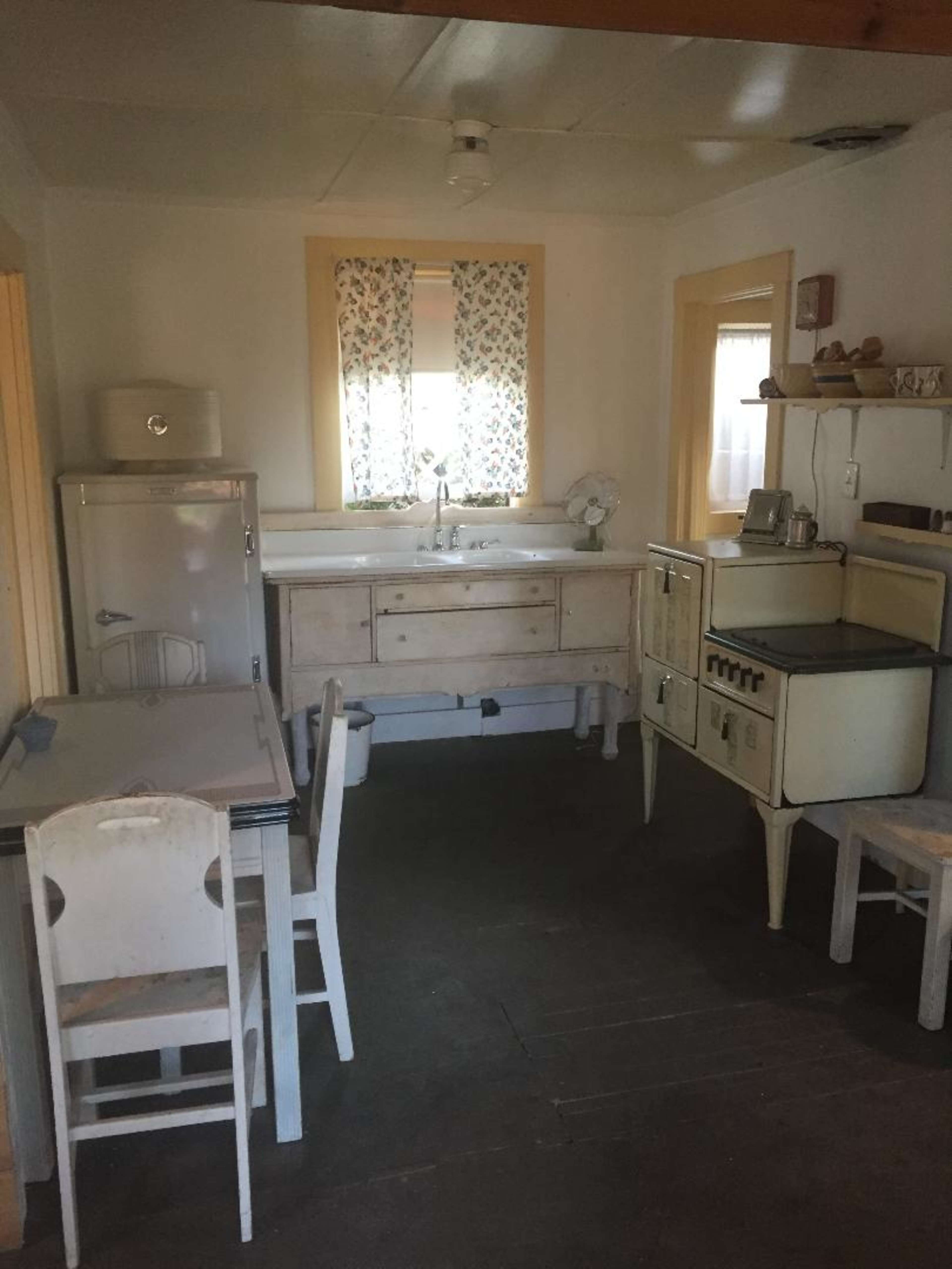 A small kitchen features a white table and chairs, a vintage refrigerator, and a sink with a window adorned with floral curtains.
