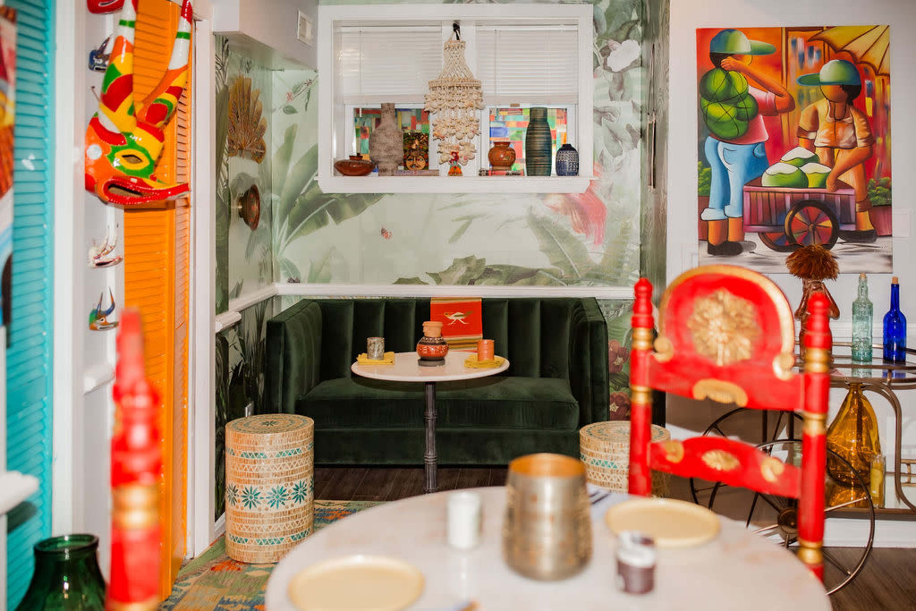 The image shows a colorful interior of a dining area featuring a green sofa, a round table with decorative items, and vibrant wall art with a tropical theme.