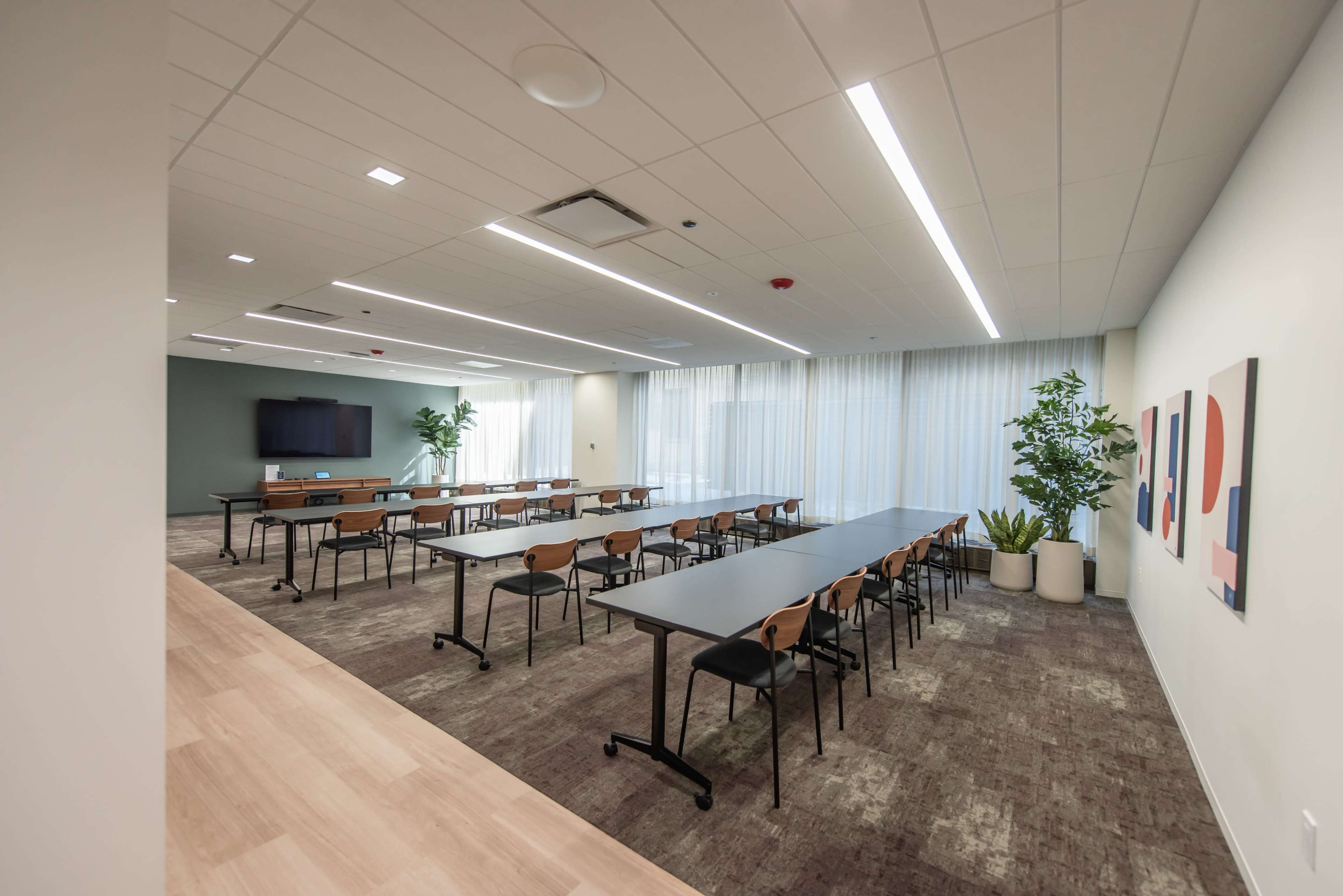 The image shows a modern conference room with several rows of tables and chairs, a large screen at one end, and potted plants near the windows.