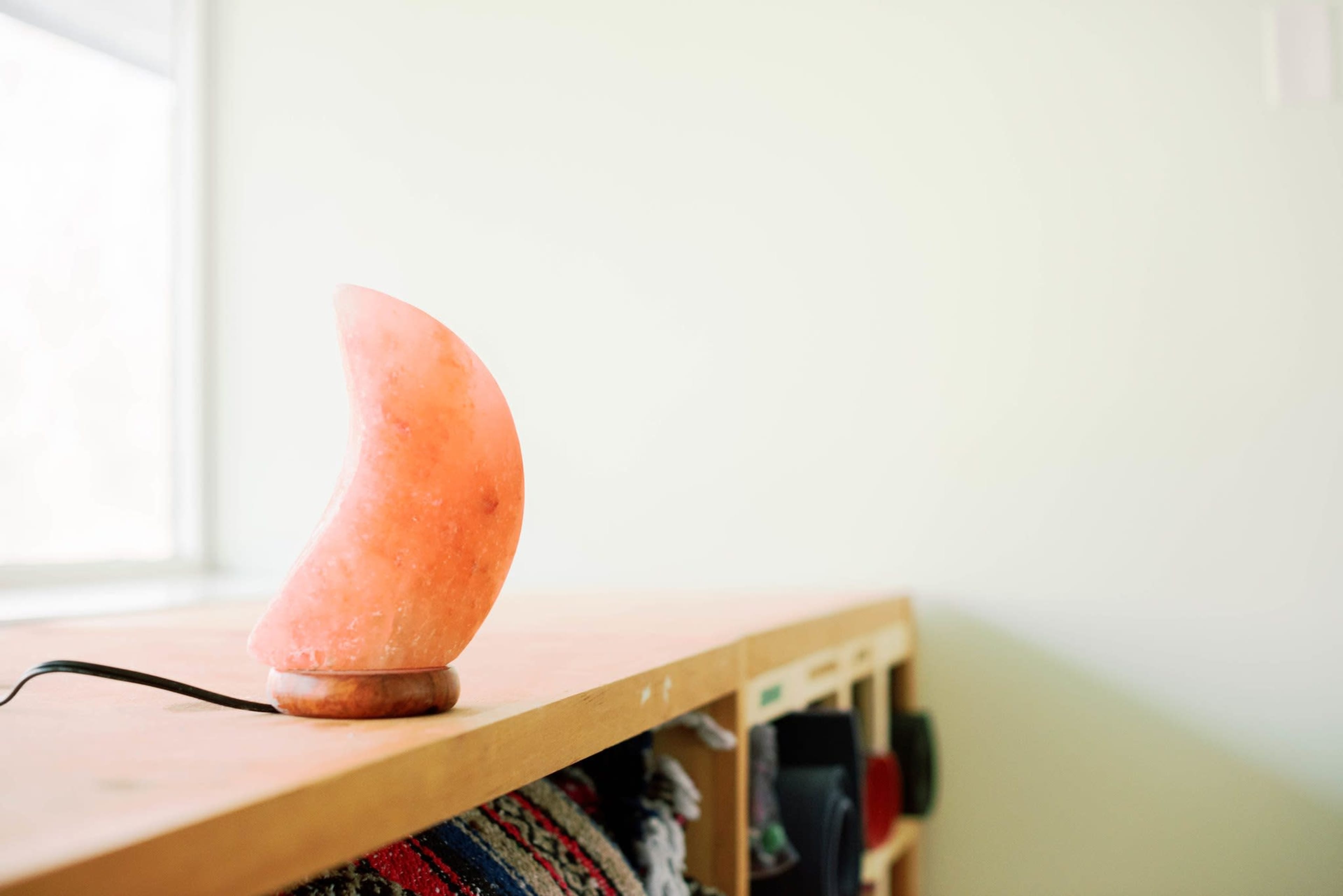 A pink salt lamp is placed on a wooden shelf next to a window in a softly lit room.