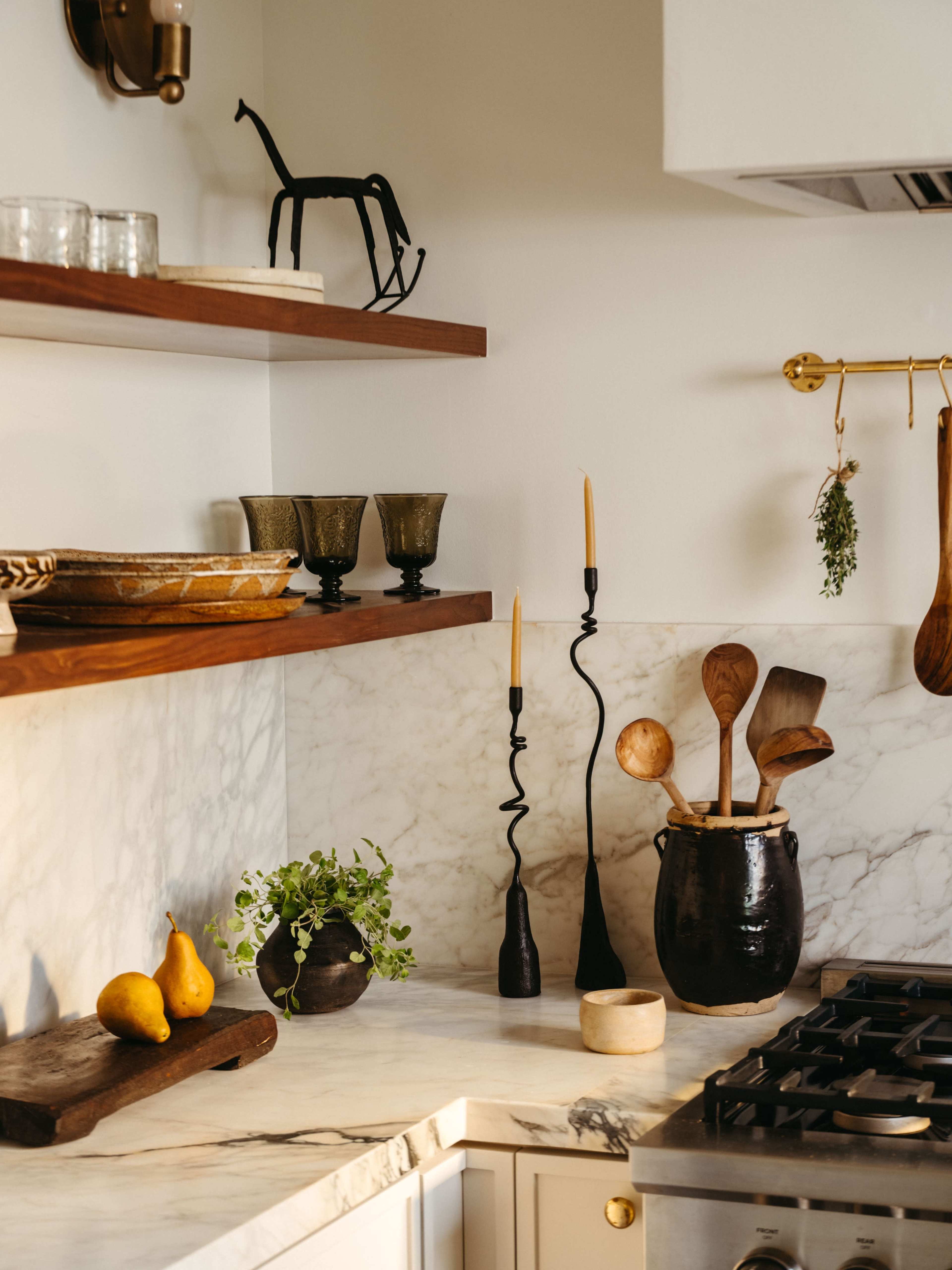 A well-organized kitchen features wooden shelves displaying glassware and tableware, with a marble countertop adorned with plants, candles, and cooking utensils.