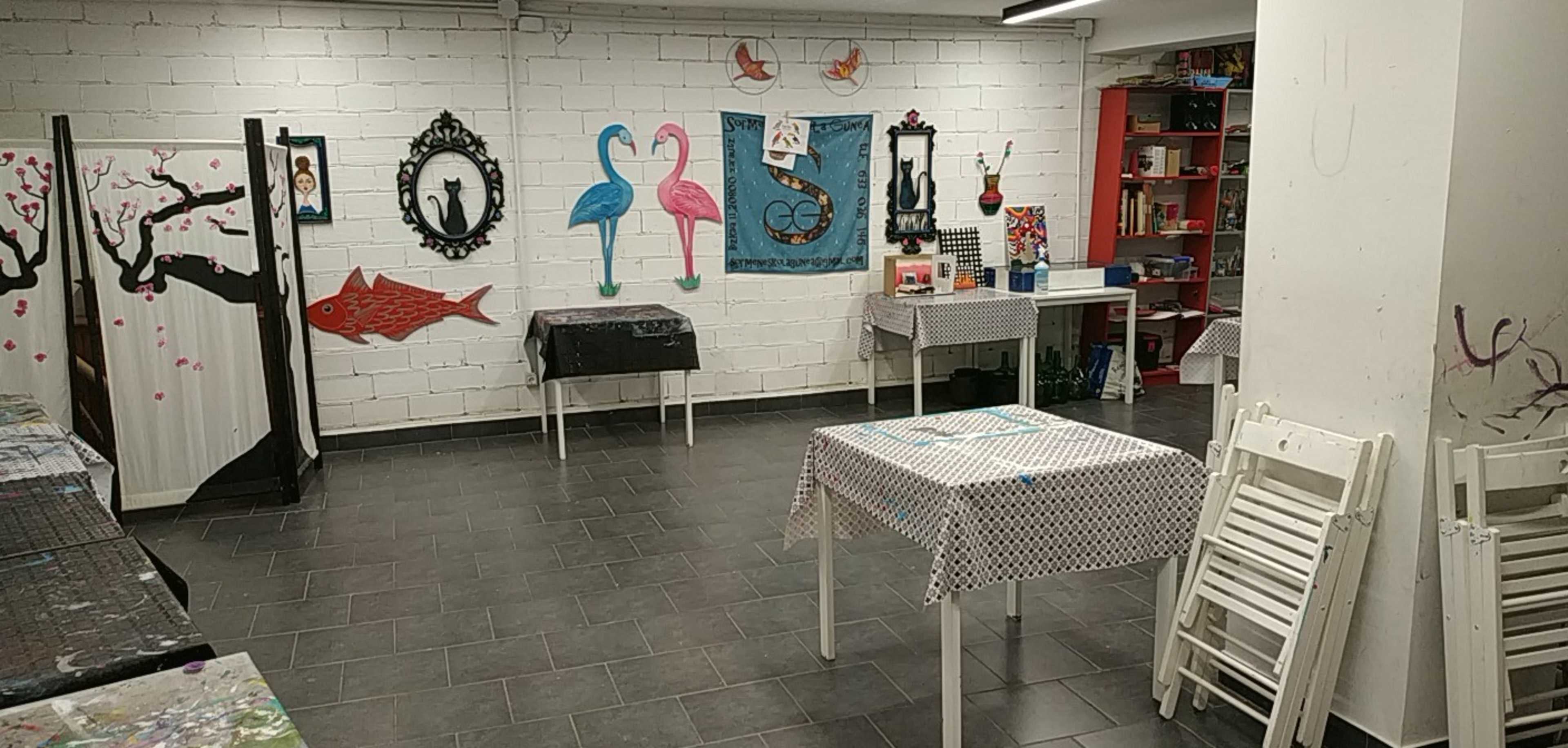 The image shows an art studio with several tables covered in cloth, decorated walls featuring various colorful art pieces, and shelves filled with art supplies.