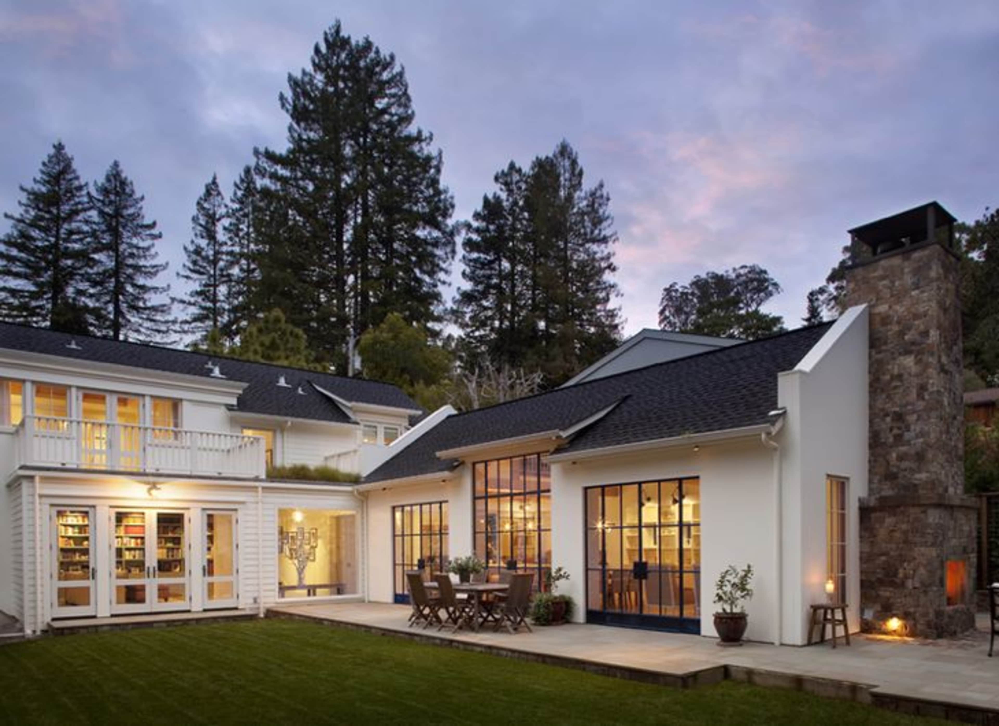 A modern two-story house with large windows, a stone chimney, and a wooden patio surrounded by trees.