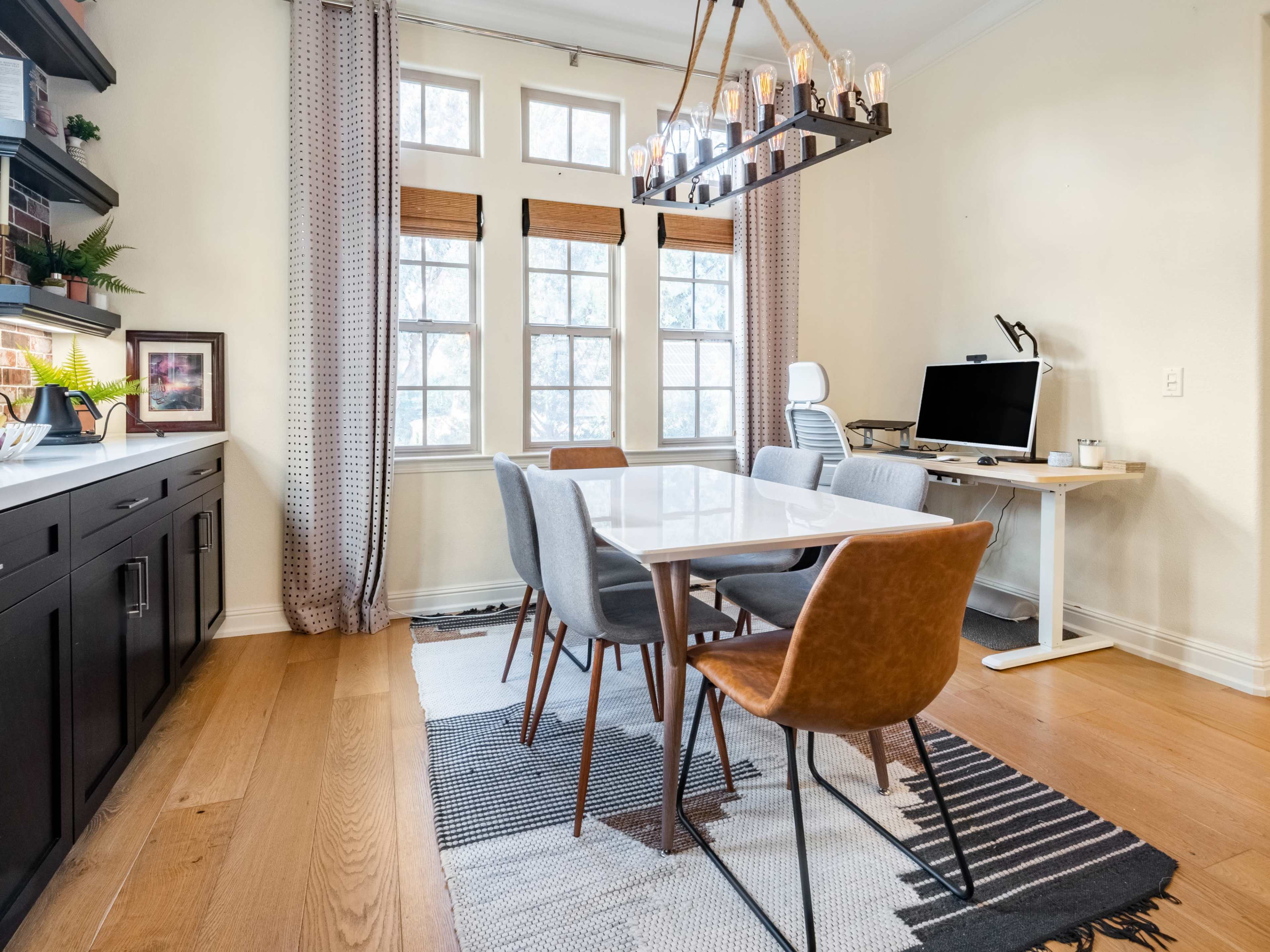 A modern dining area features a long white table surrounded by various chairs, with a large window letting in natural light and a computer workstation in the corner.