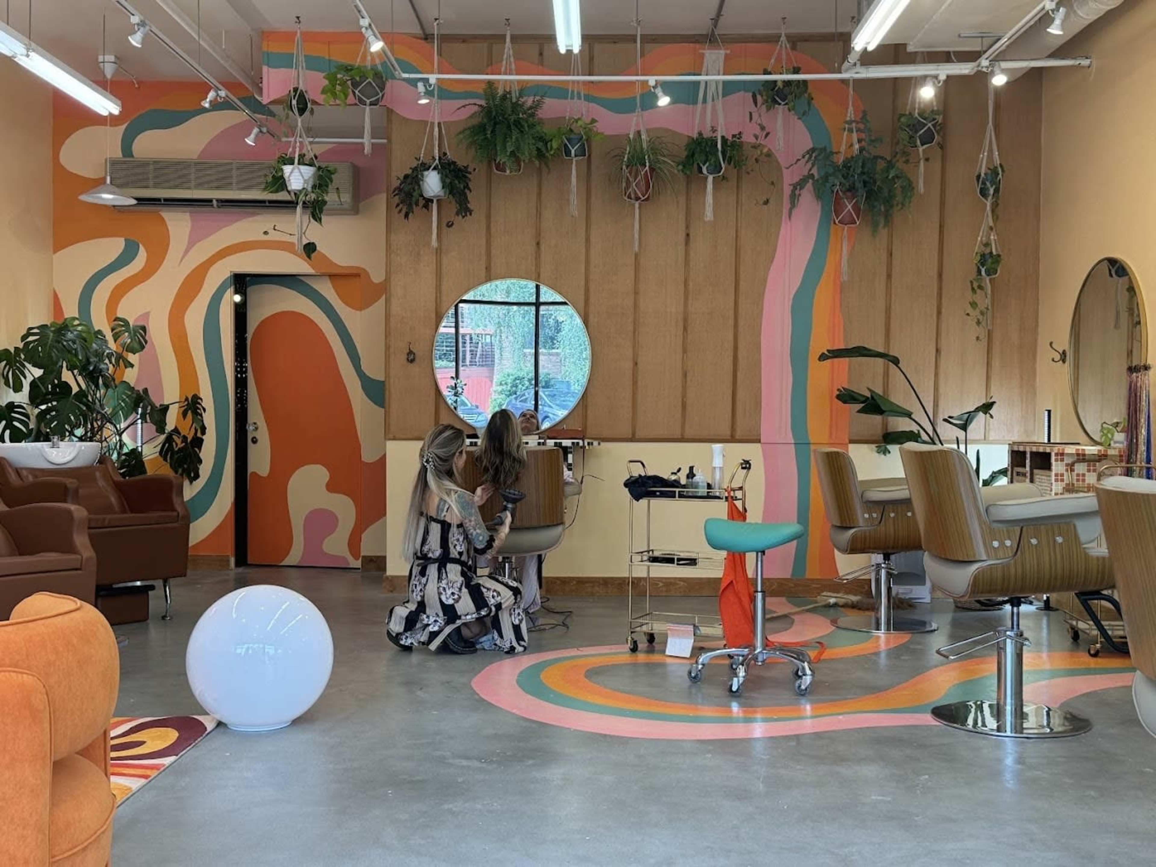 A hair salon interior features colorful abstract wall designs, plants hanging from the ceiling, and two people engaged in a hair treatment near a circular window.