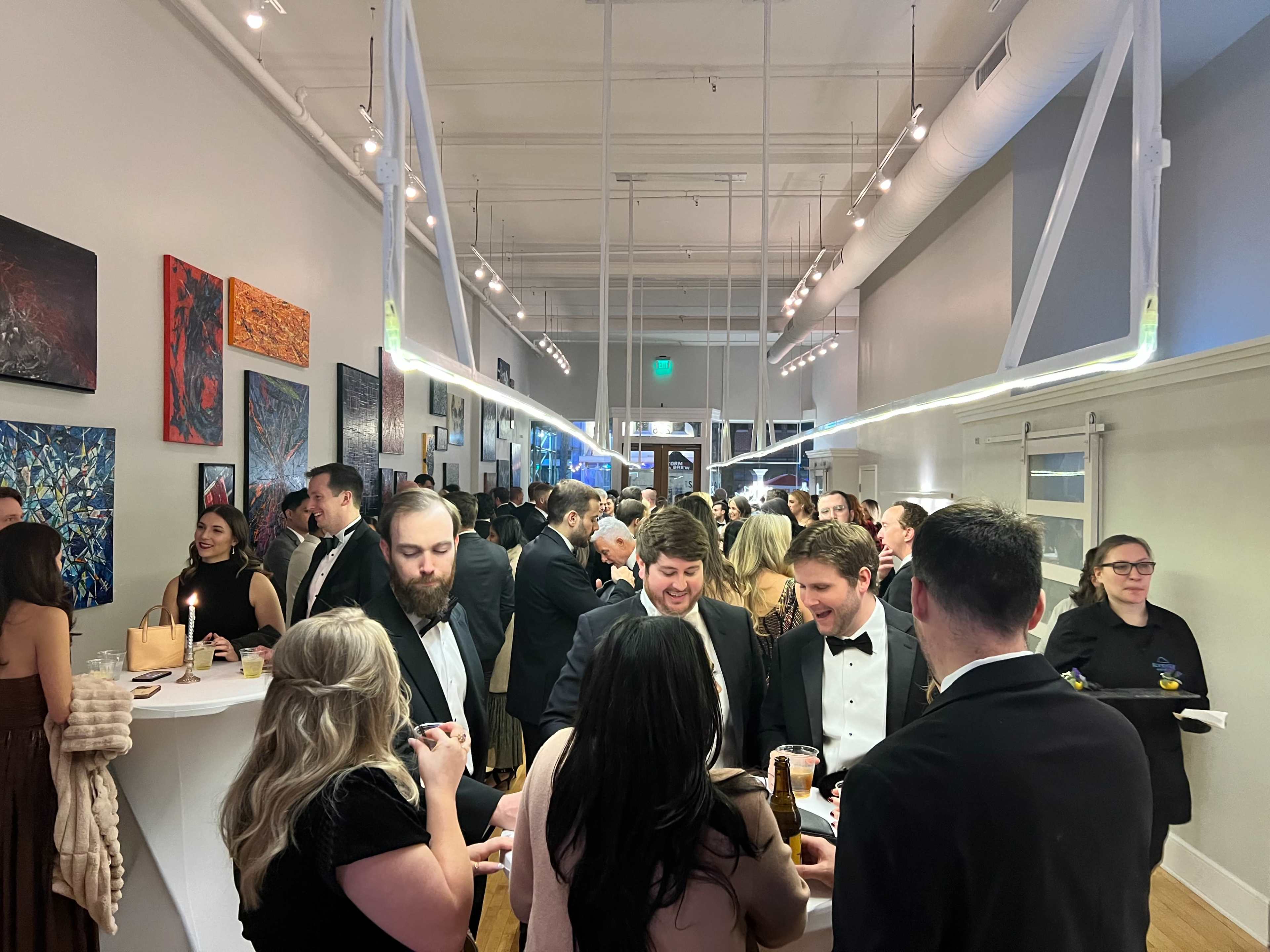 A crowded indoor venue featuring guests in formal attire engaged in conversation and socializing amidst colorful artwork on the walls.