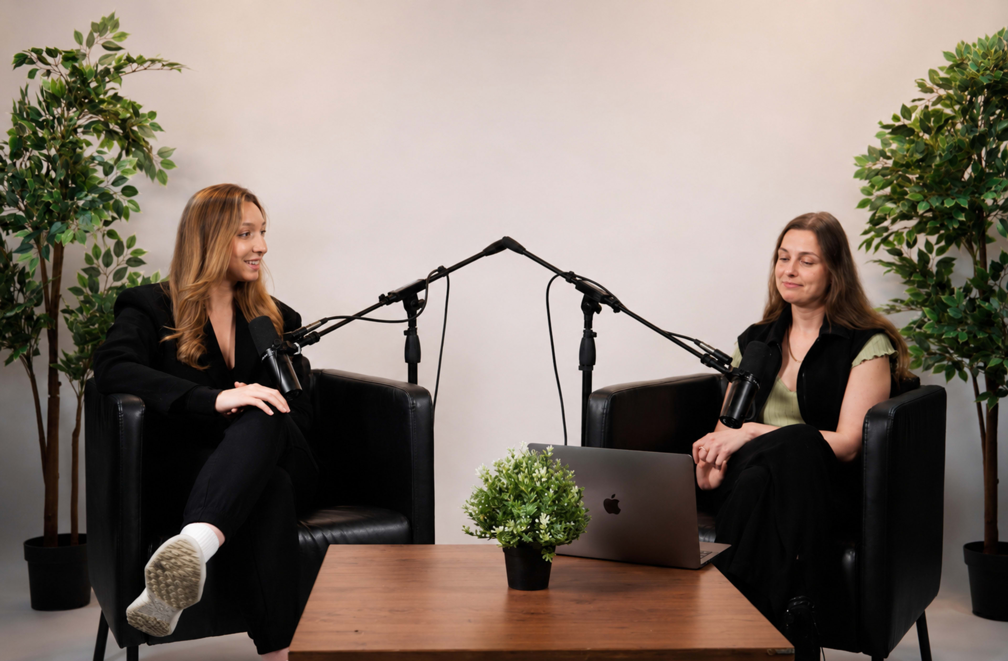 Two women are seated in black chairs across from each other, with microphones positioned between them and a laptop on a wooden table, surrounded by potted plants.