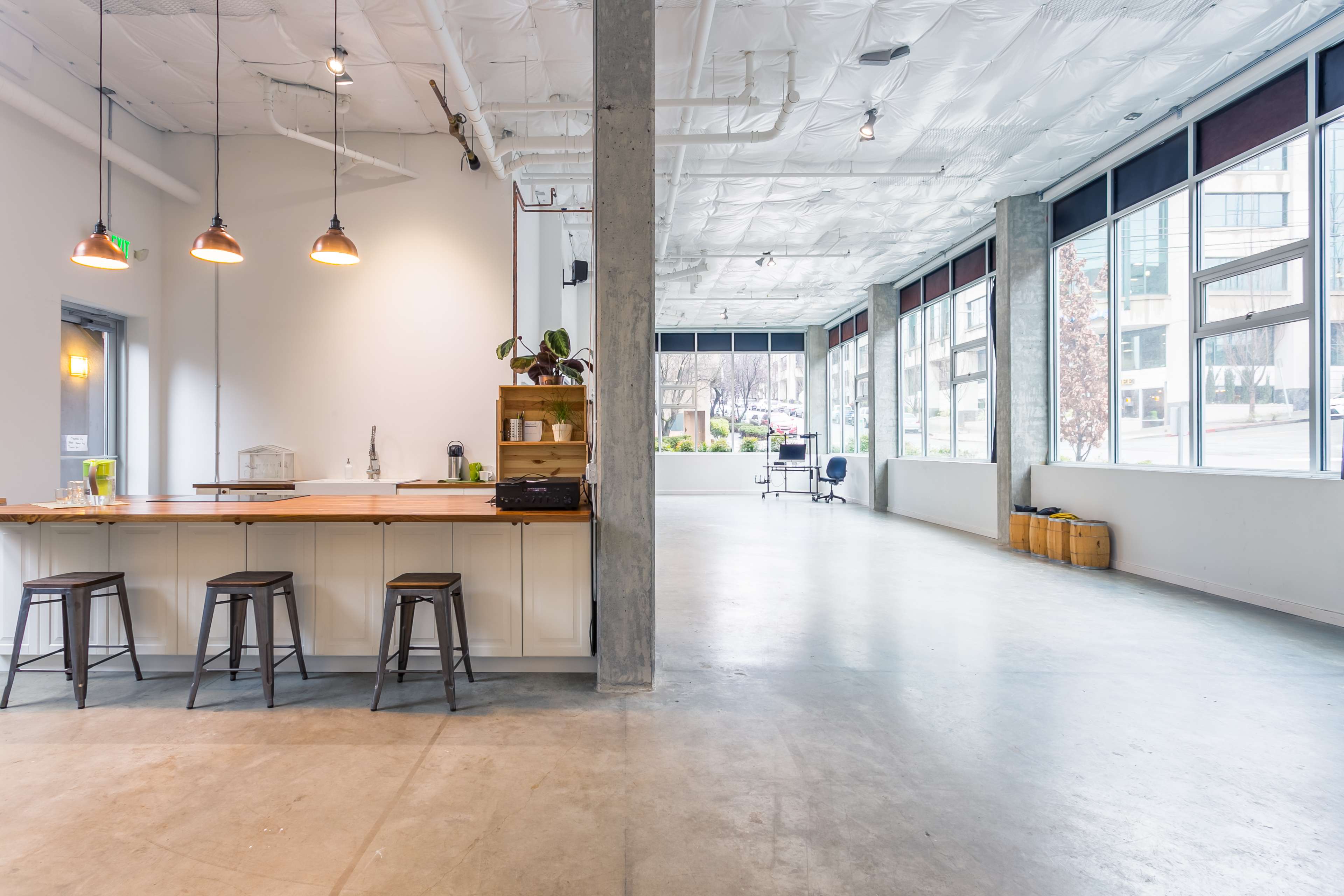 An airy, modern workspace with a kitchen island, bar stools, large windows, and a polished concrete floor.