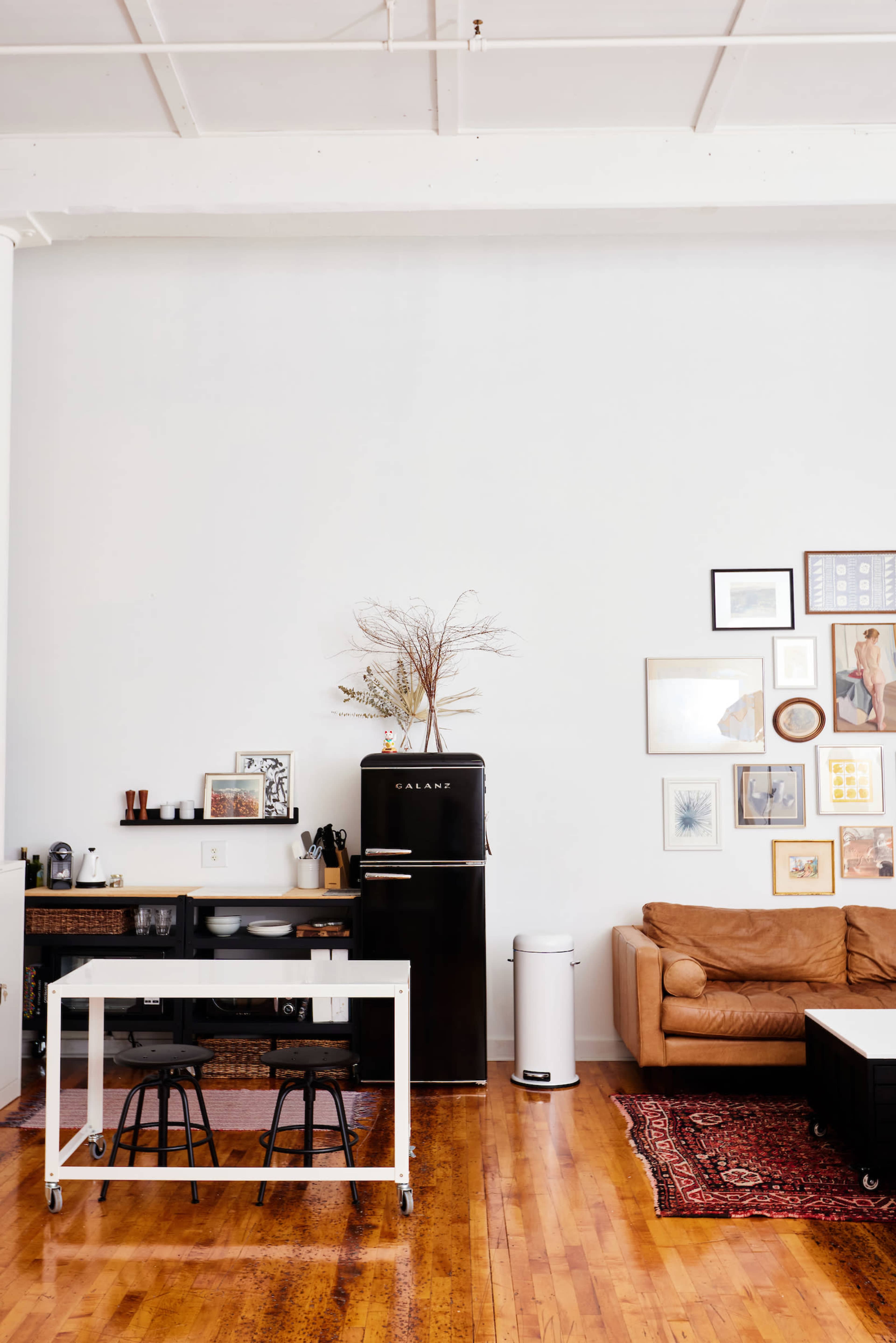 A modern, minimalist kitchen and living area features a black refrigerator, a wooden dining table with two stools, and a brown couch against a wall adorned with framed artwork.