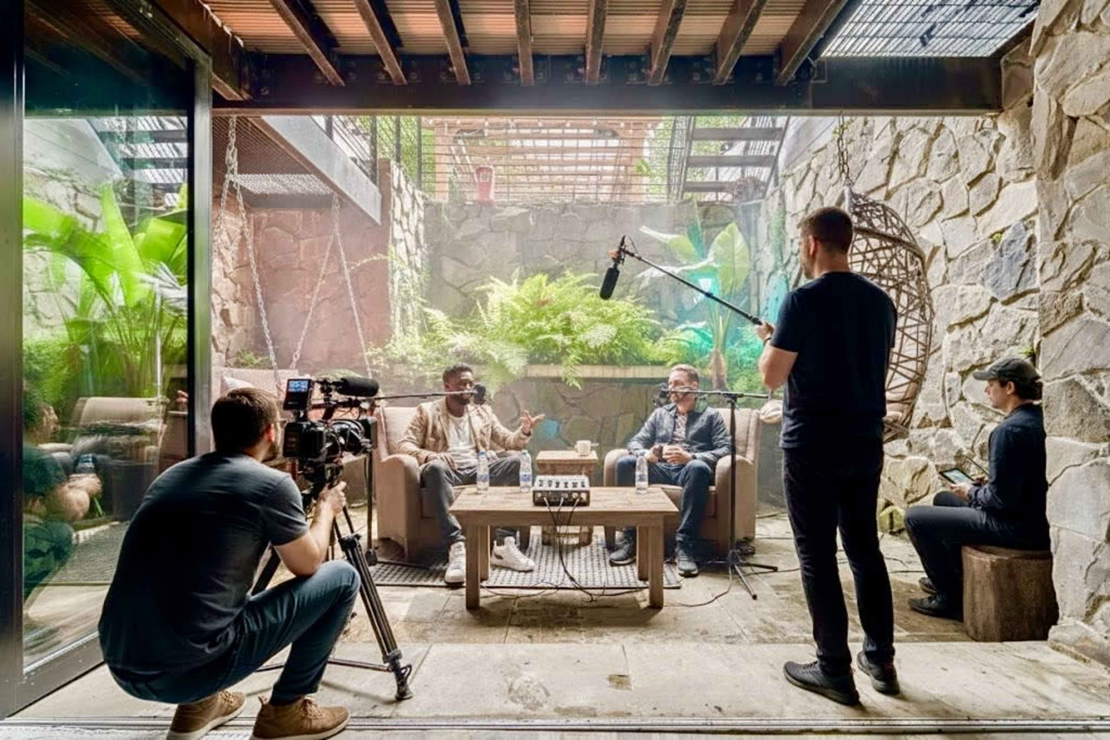 Two men are seated on a couch in a modern indoor setting with plants and stone walls, while a crew member films the conversation with a camera and microphone.