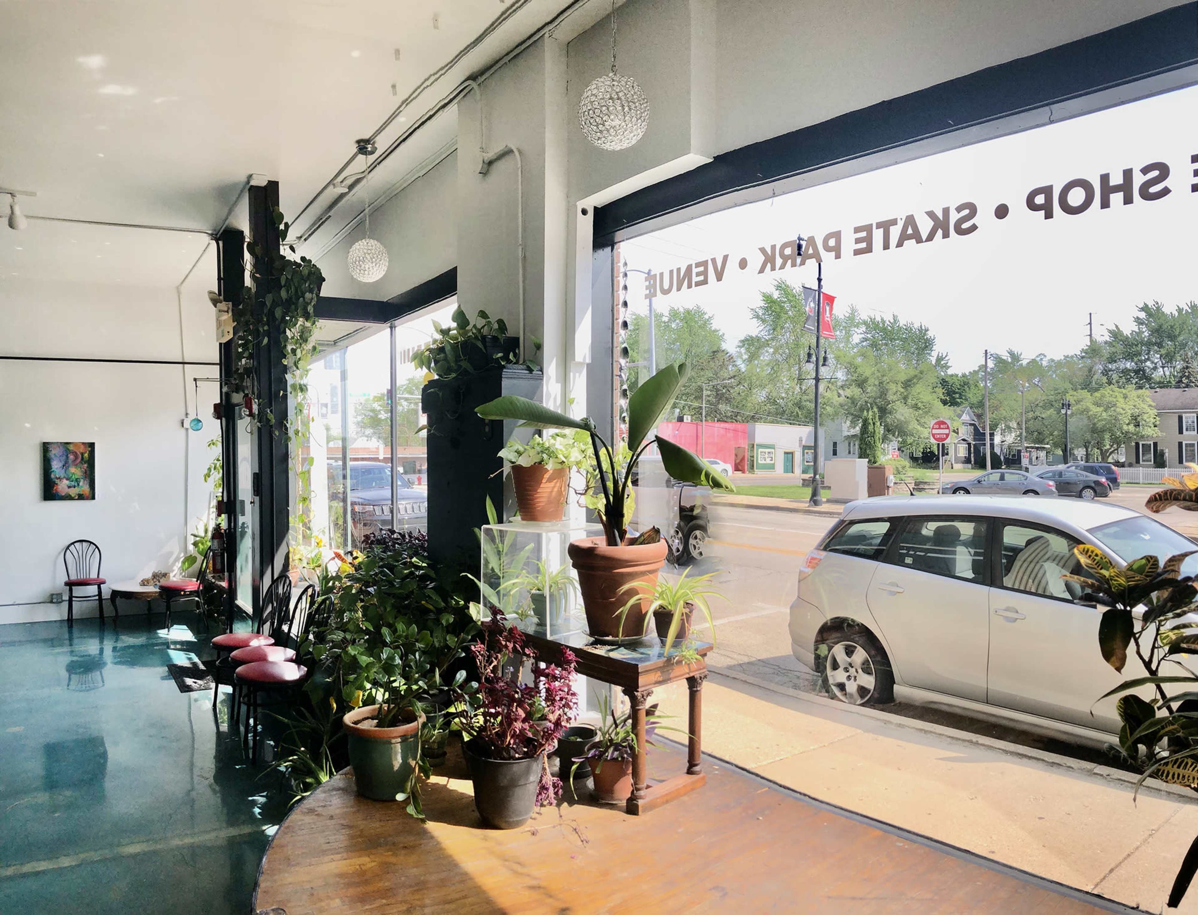A bright interior of a shop features plants and seating by a large window overlooking a busy street.