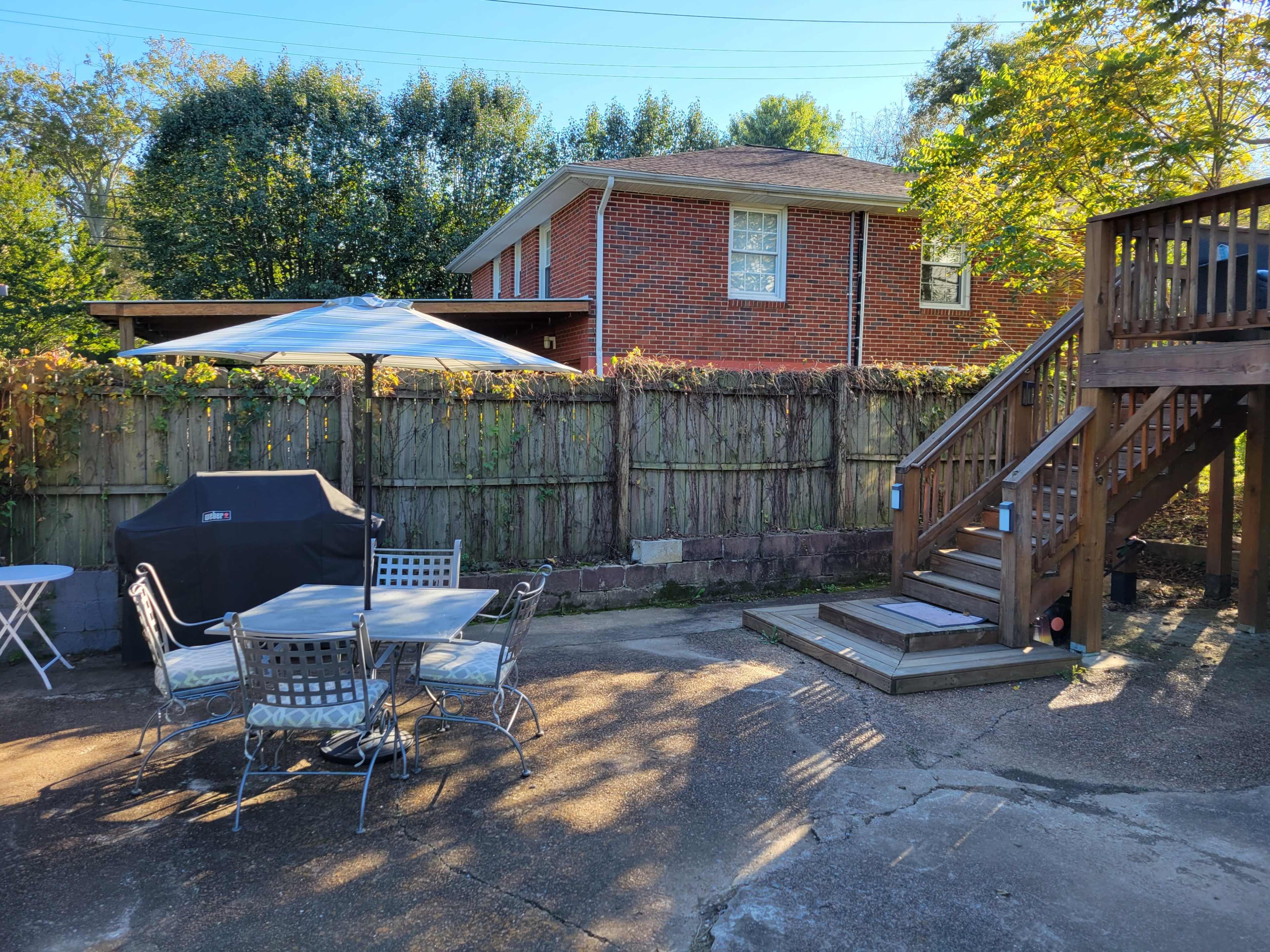 A backyard features a seating area with a table and chairs, a grill under an umbrella, and a wooden deck leading to a house.