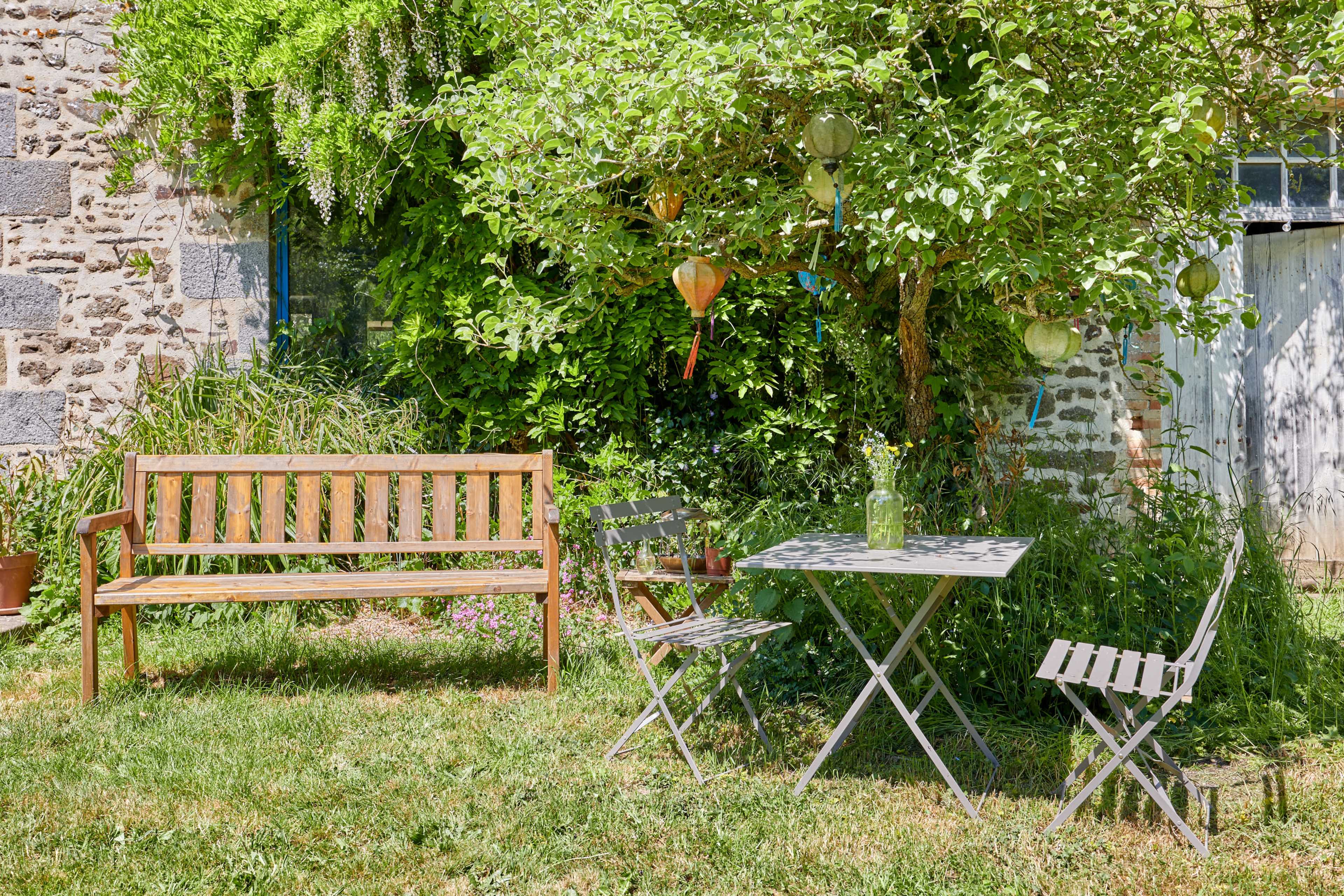 A wooden bench and a small folding table with chairs sit beneath a lush tree in a garden setting.