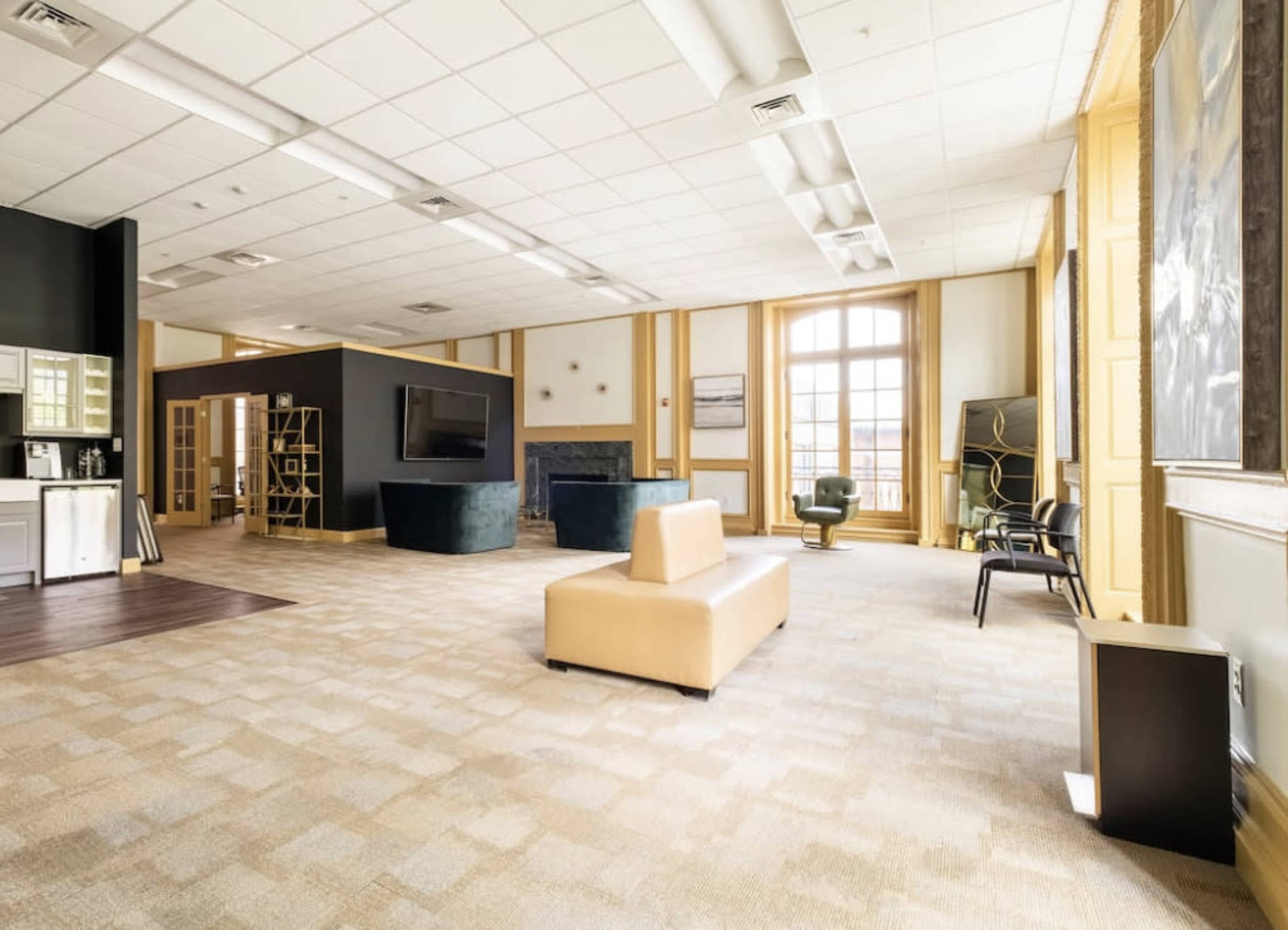 A spacious, modern office area featuring light-colored walls, large windows, and a mix of seating arrangements, including a sofa and chairs.