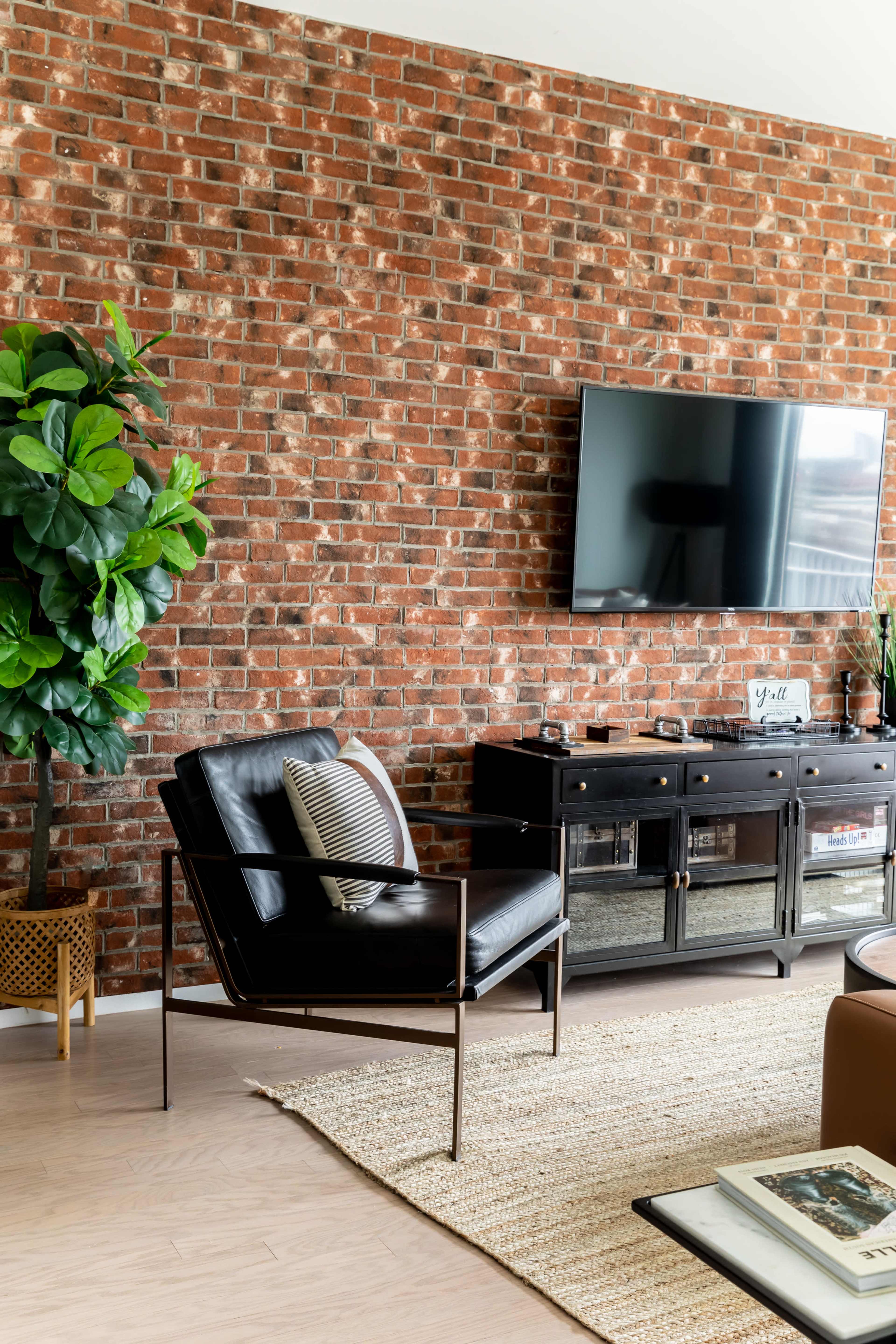 The image depicts a modern living room with a black armchair, a television mounted on a brick wall, and a potted plant beside a dark wooden console table.