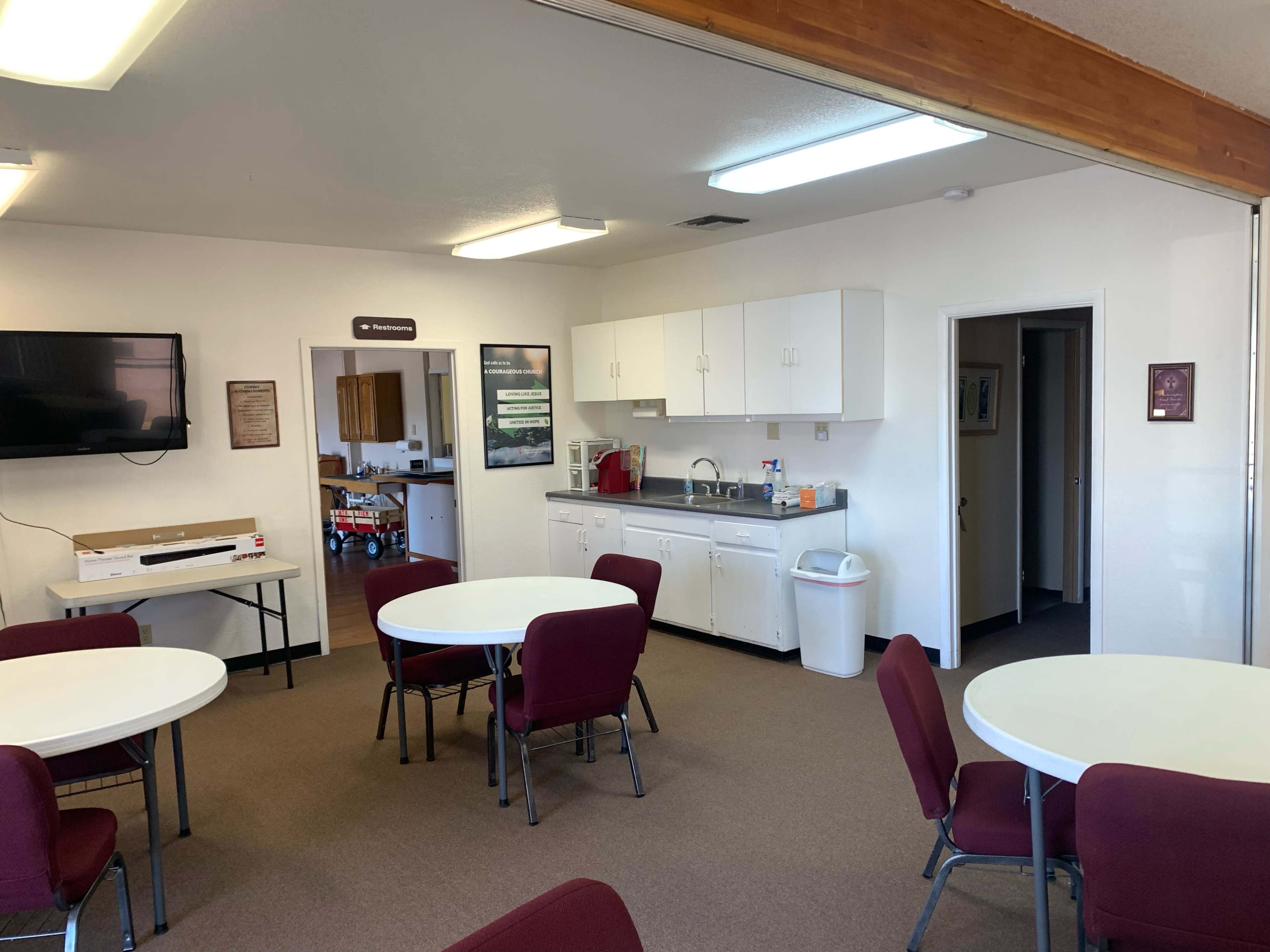 A clean, well-lit communal kitchen and dining area featuring several round tables with chairs, a sink, and a television on the wall.