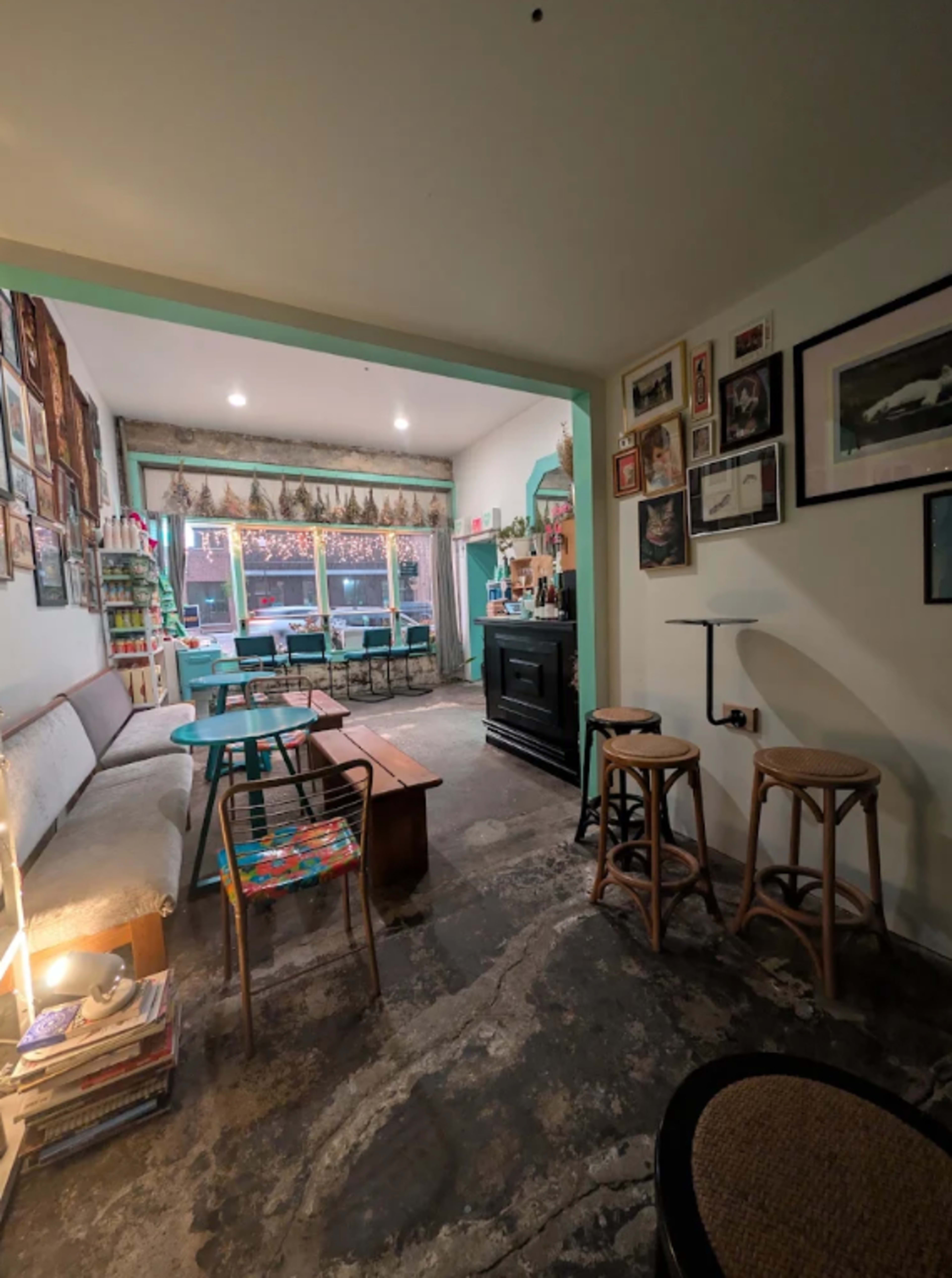The image shows a cozy cafe interior with a seating area featuring a couch, tables, and stools, adorned with artwork on the walls and a large window in the background.