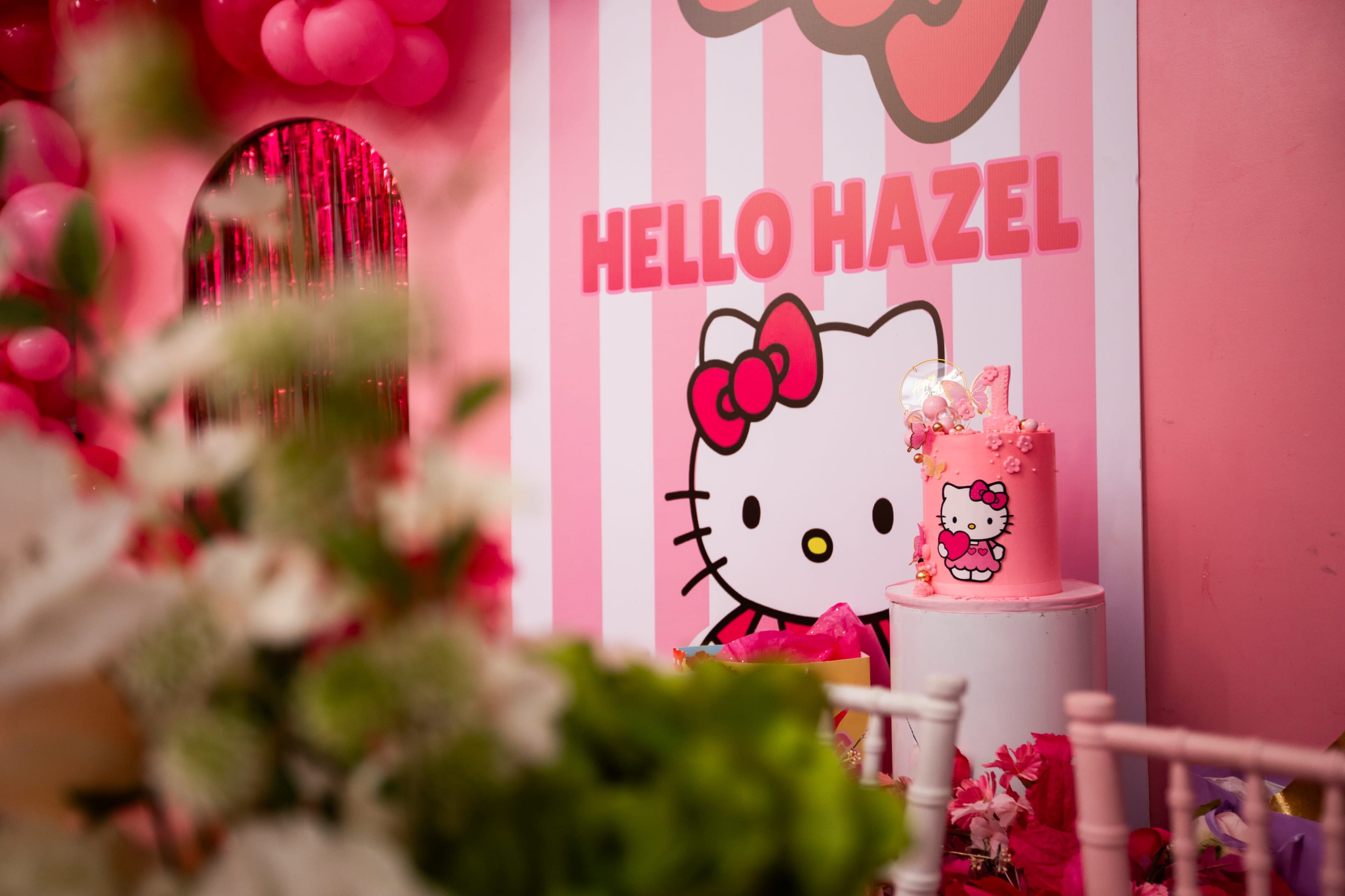 The image features a pink and white striped backdrop with a Hello Kitty design, a cake adorned with Hello Kitty elements, and various pink decorations.