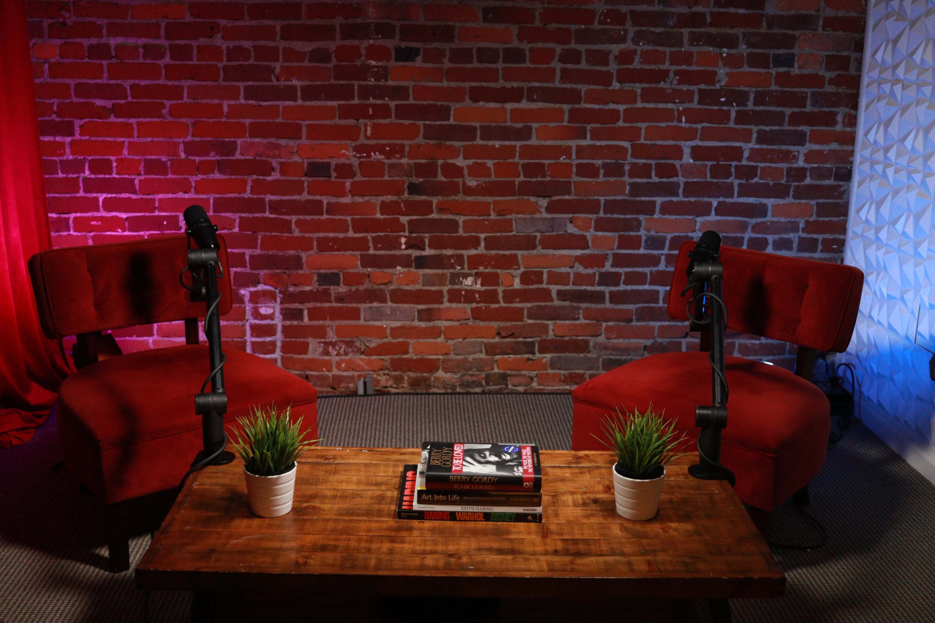 Two red chairs with microphones positioned at a wooden table, set against a brick wall with colored lighting.