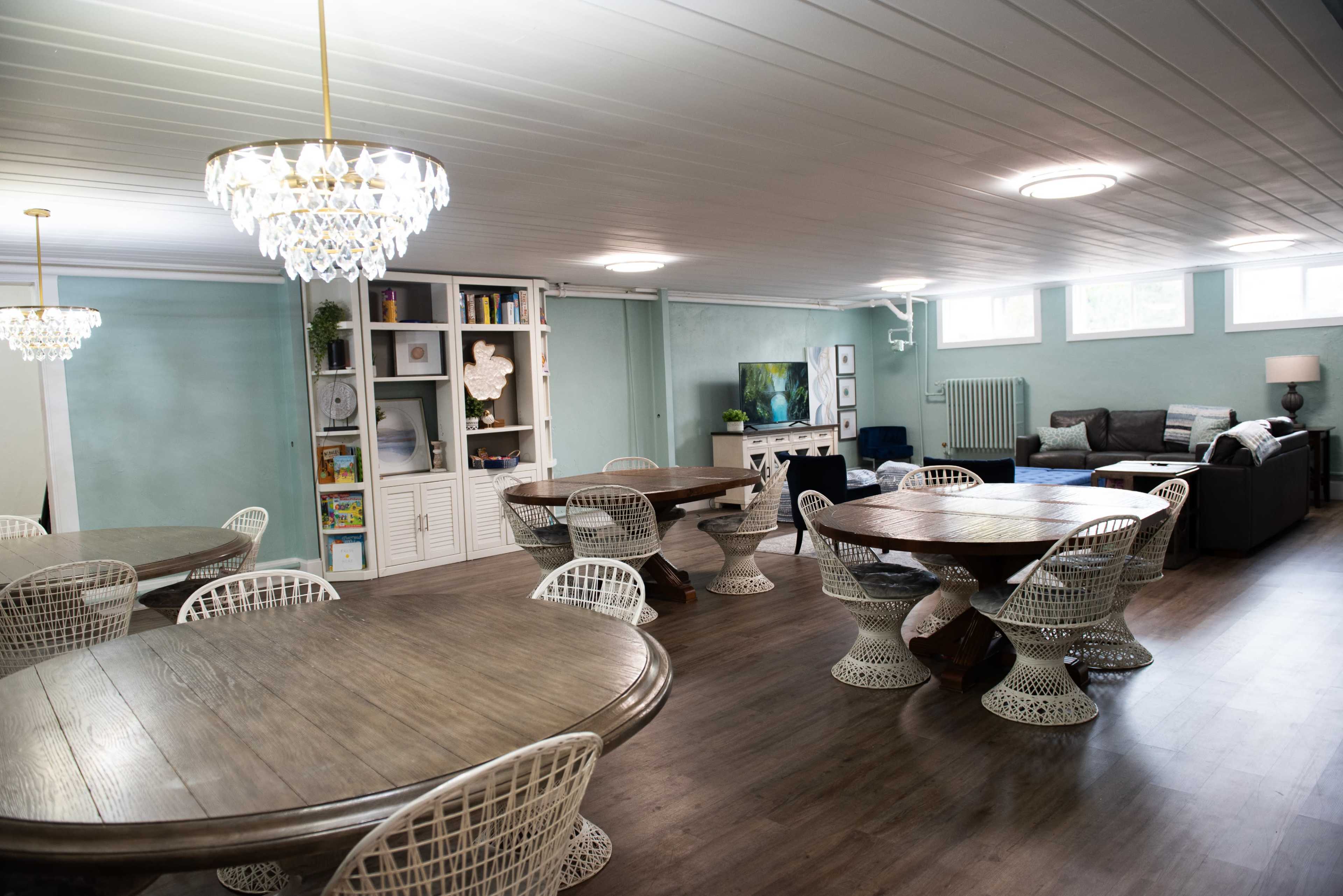 The image shows a spacious room featuring multiple round tables surrounded by wicker chairs, with a seating area containing a couch and a television in the background.